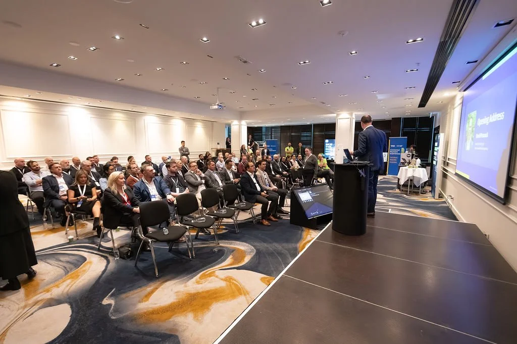A man in a suit giving a presentation on stage to an audience at a conference in a large, modern room with a projector and screen.