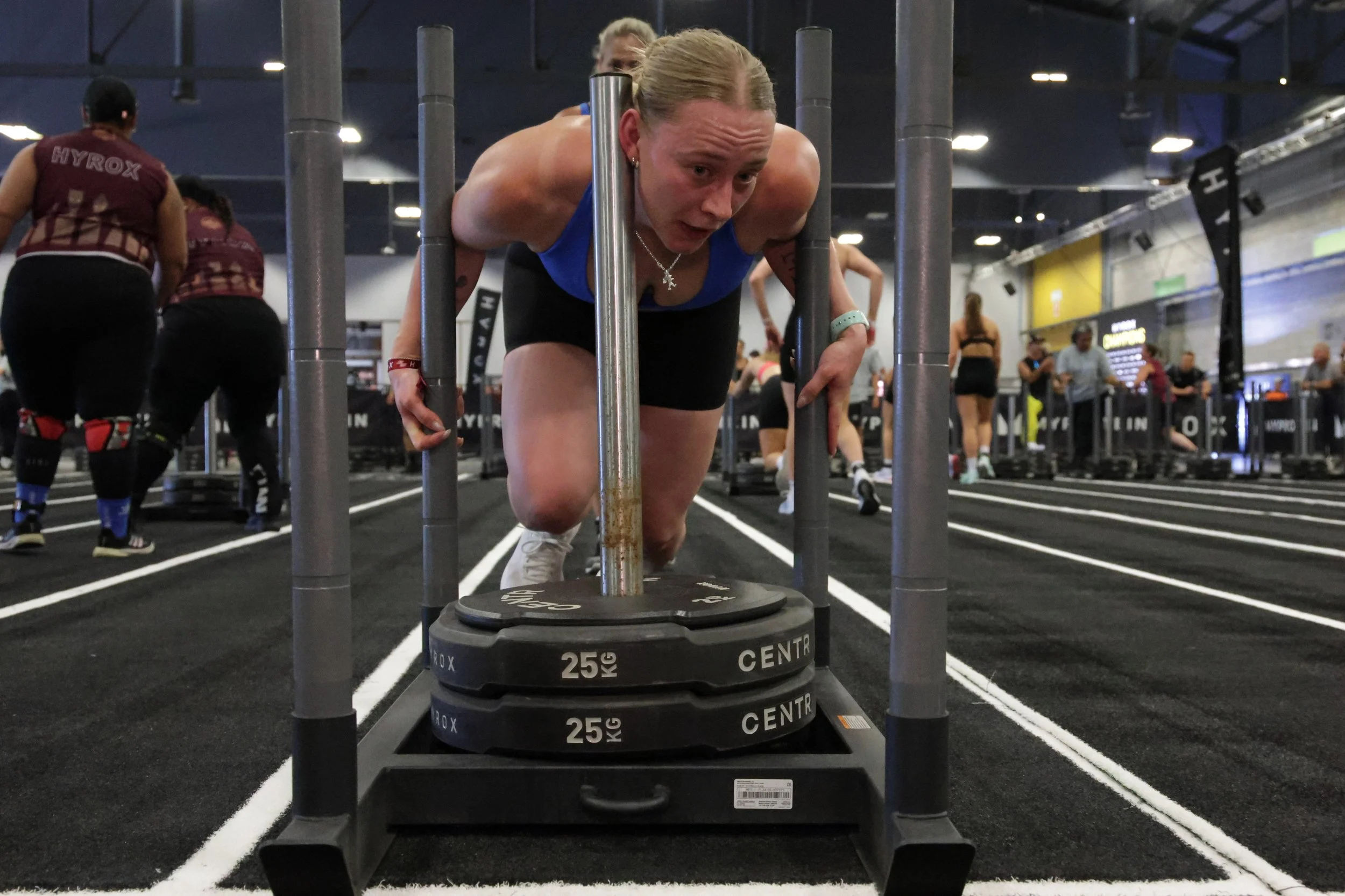 A woman in athletic gear pushing weights on a black gym floor with white court lines, surrounded by other gym-goers.