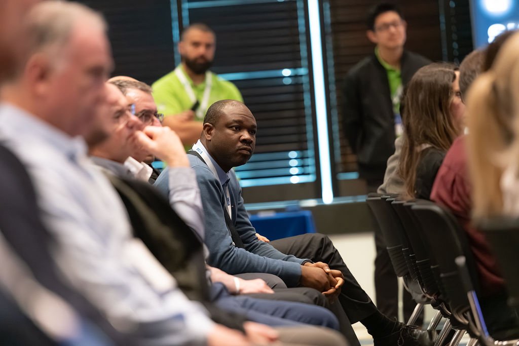 A group of diverse people attending a conference or seminar, seated and listening attentively, with some standing in the background.