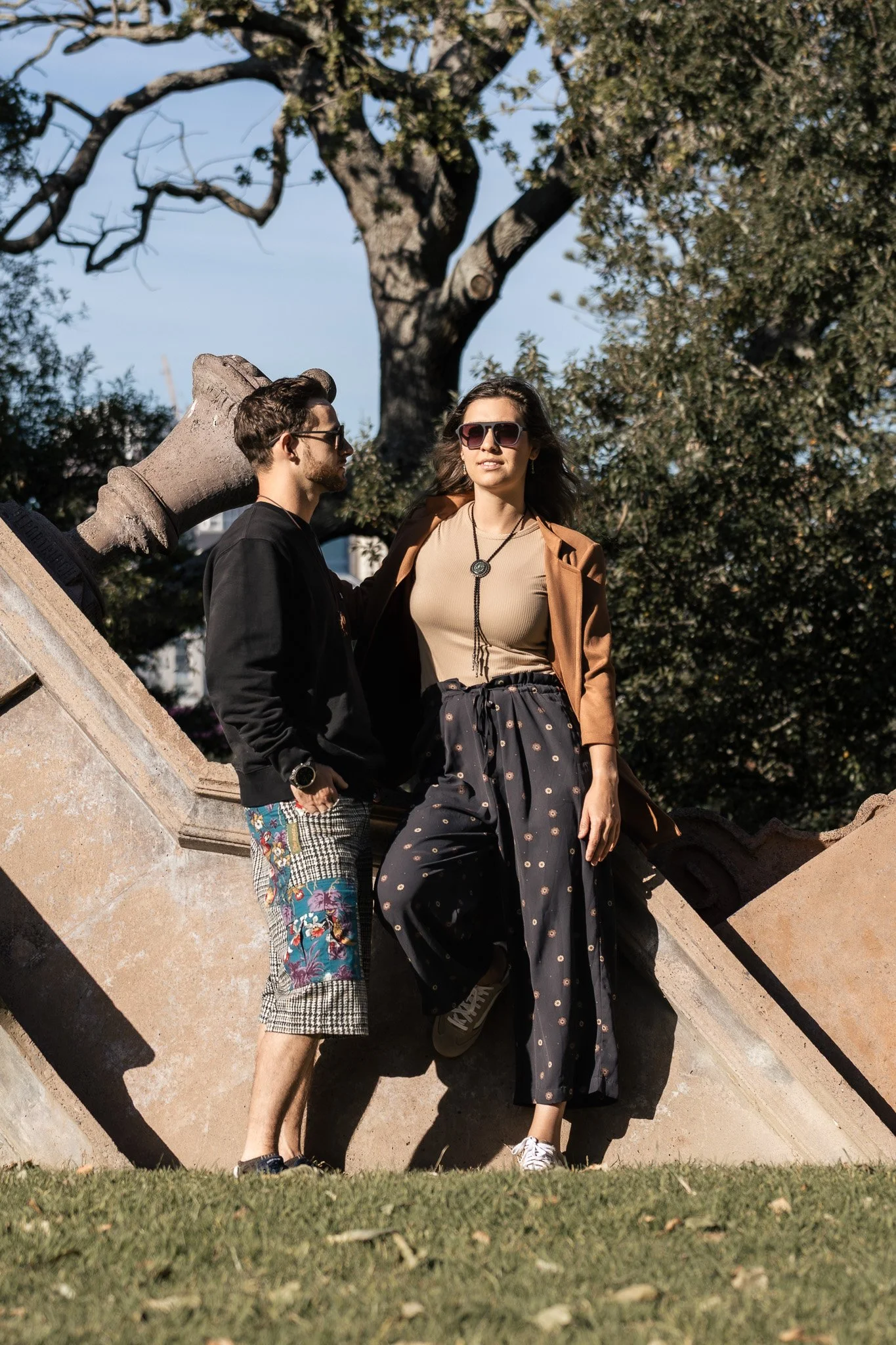 A young man and woman standing on a concrete staircase outdoors, with a large tree in the background. The woman wears sunglasses, a tan jacket, and a black skirt with pattern, while the man wears sunglasses, a black jacket, and colorful patterned sho