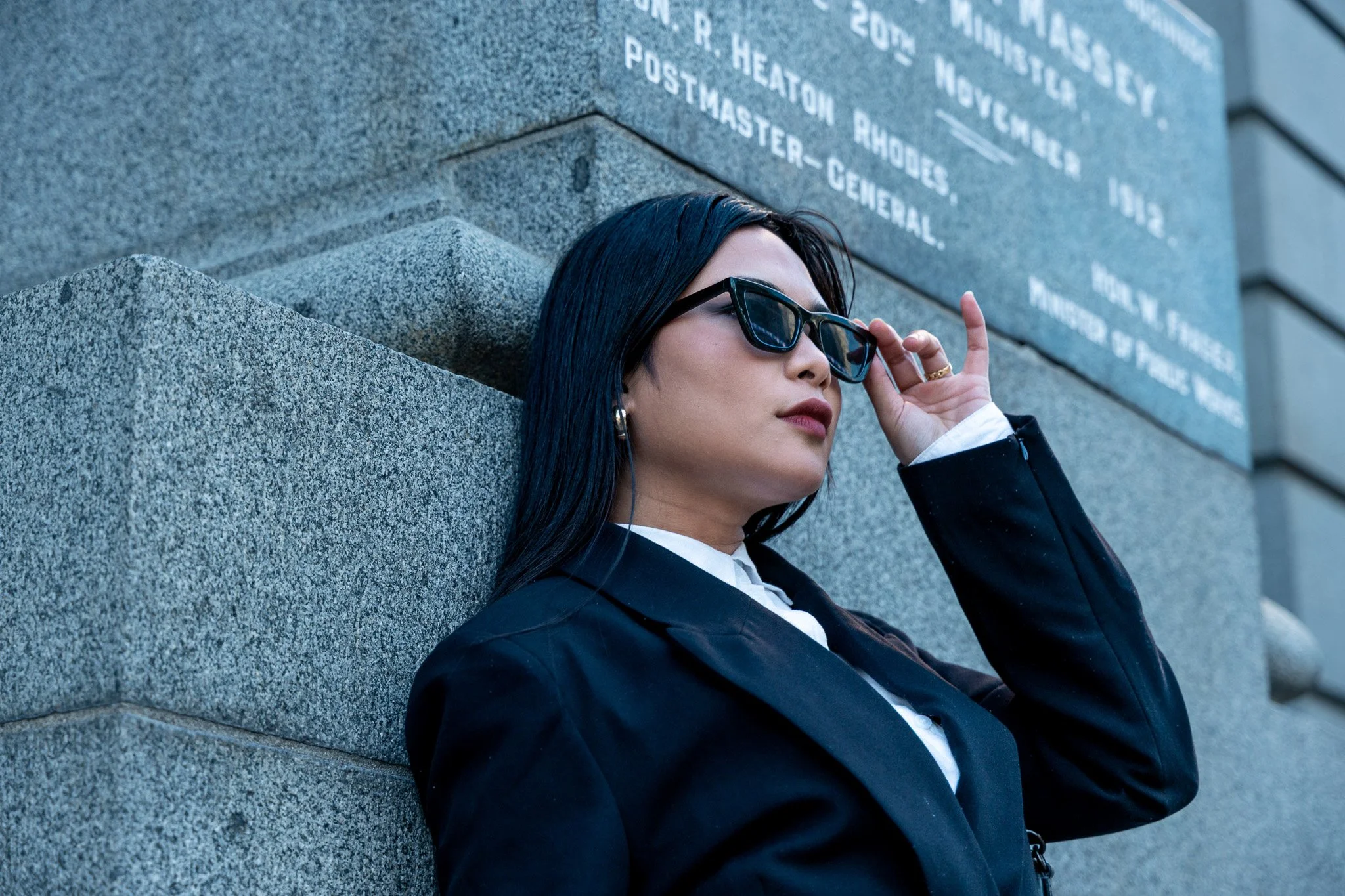 A woman with long black hair and sunglasses leaning against a stone monument with engraved text.
