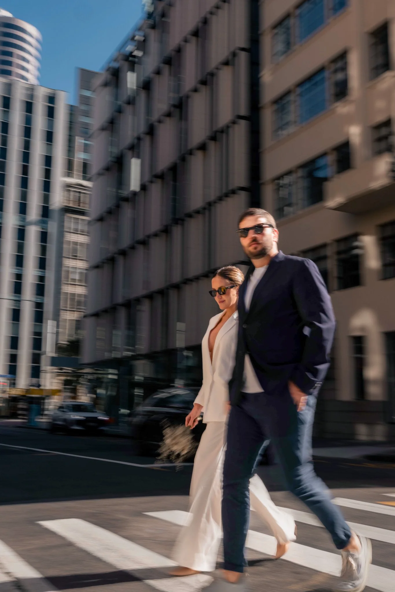 A man and woman crossing a city street at a crosswalk with modern buildings in the background.