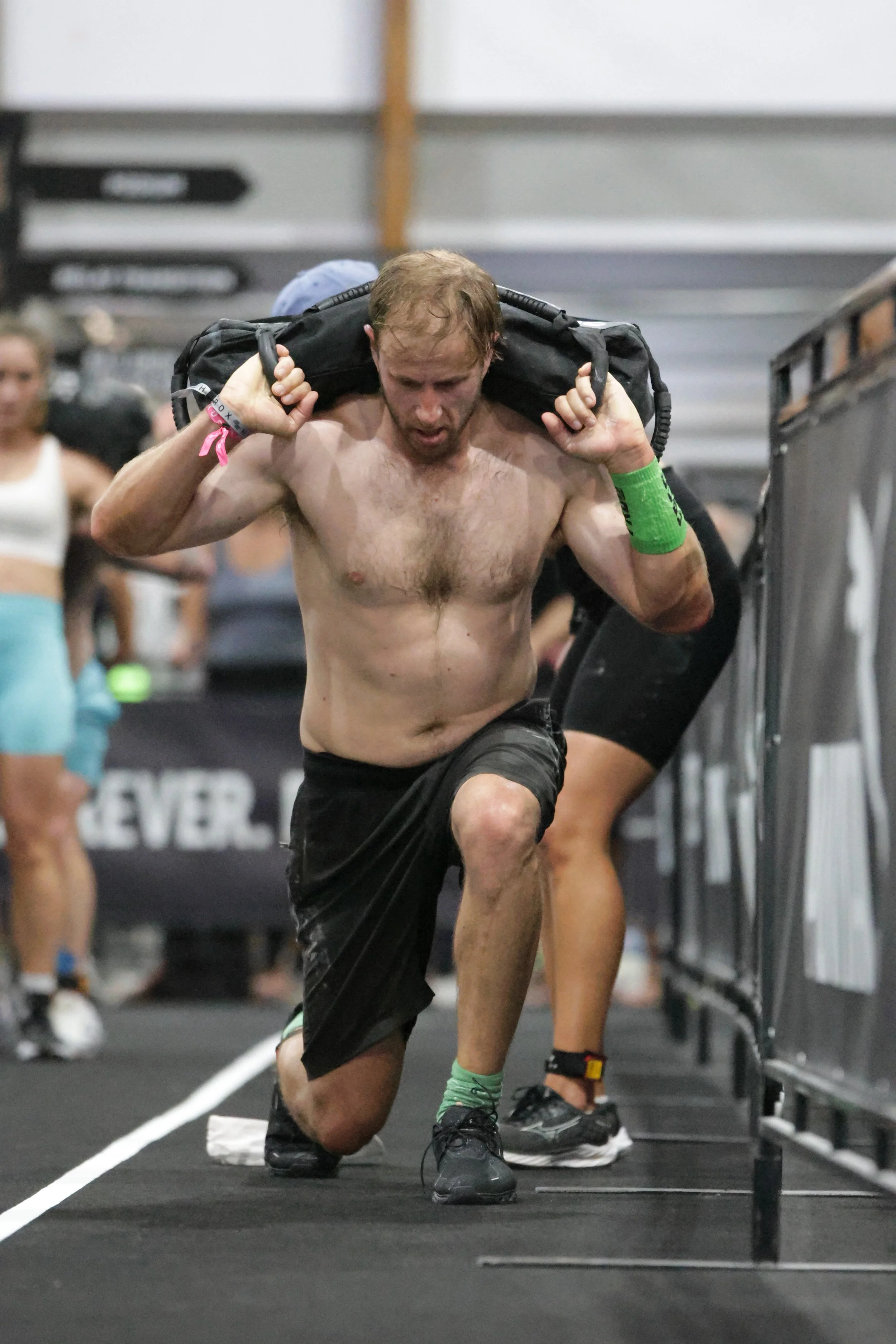 A shirtless man with a green wristband, black shorts, and black sneakers carrying a backpack on his shoulders, running on a treadmill in a gym. Behind him, a woman in a white top and blue shorts is visible, with other gym members blurred in the backg