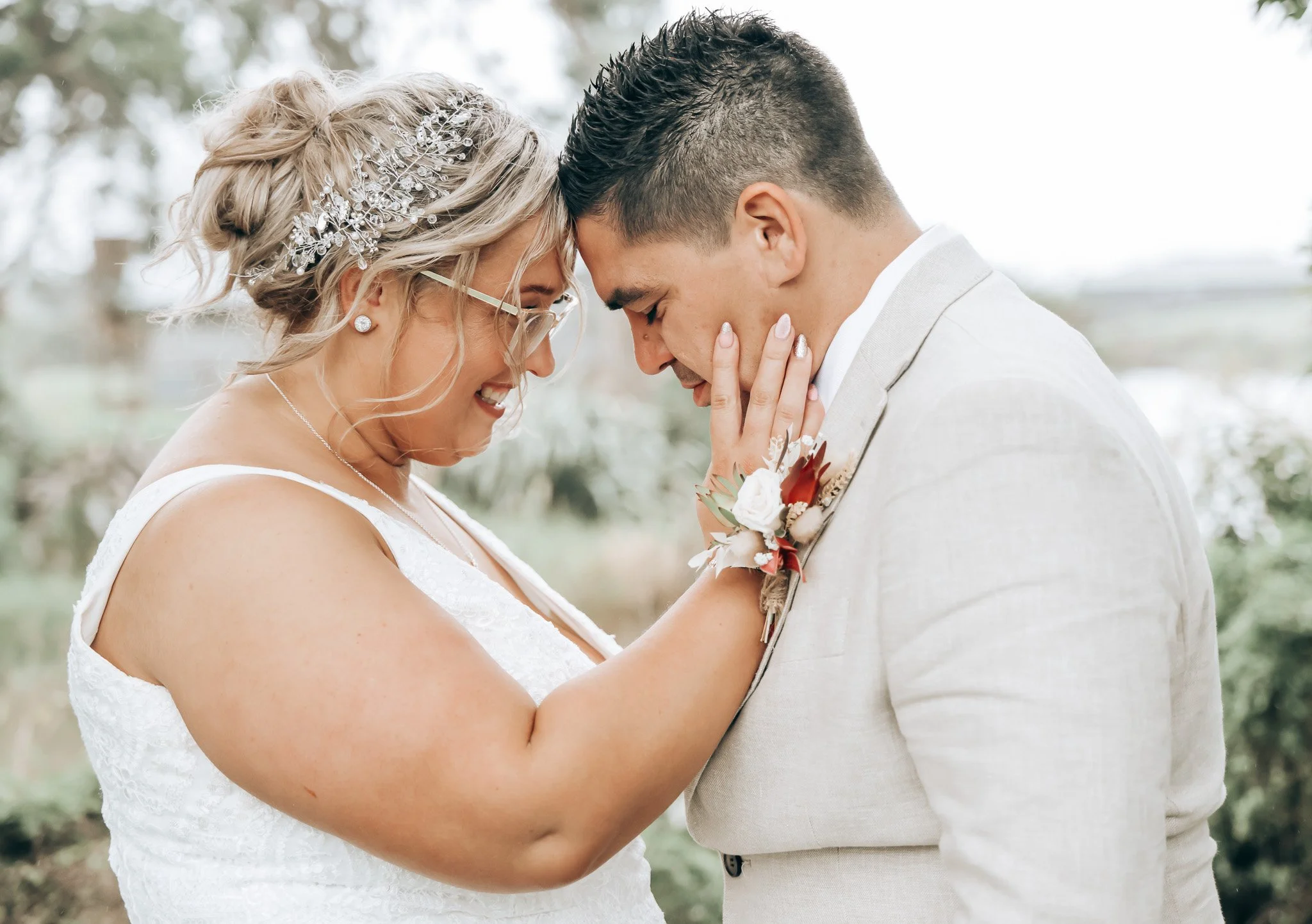 A bride and groom standing close together outdoors, touching foreheads and smiling with eyes closed. The bride has blonde hair styled in loose waves with a decorative hairpiece, glasses, and is wearing a white lace wedding dress. The groom has short 