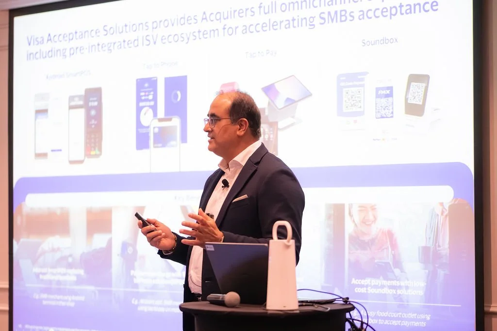 A man in a dark suit and glasses giving a presentation in front of a large screen displaying various images of smartphones and QR codes related to Visa acceptance solutions.