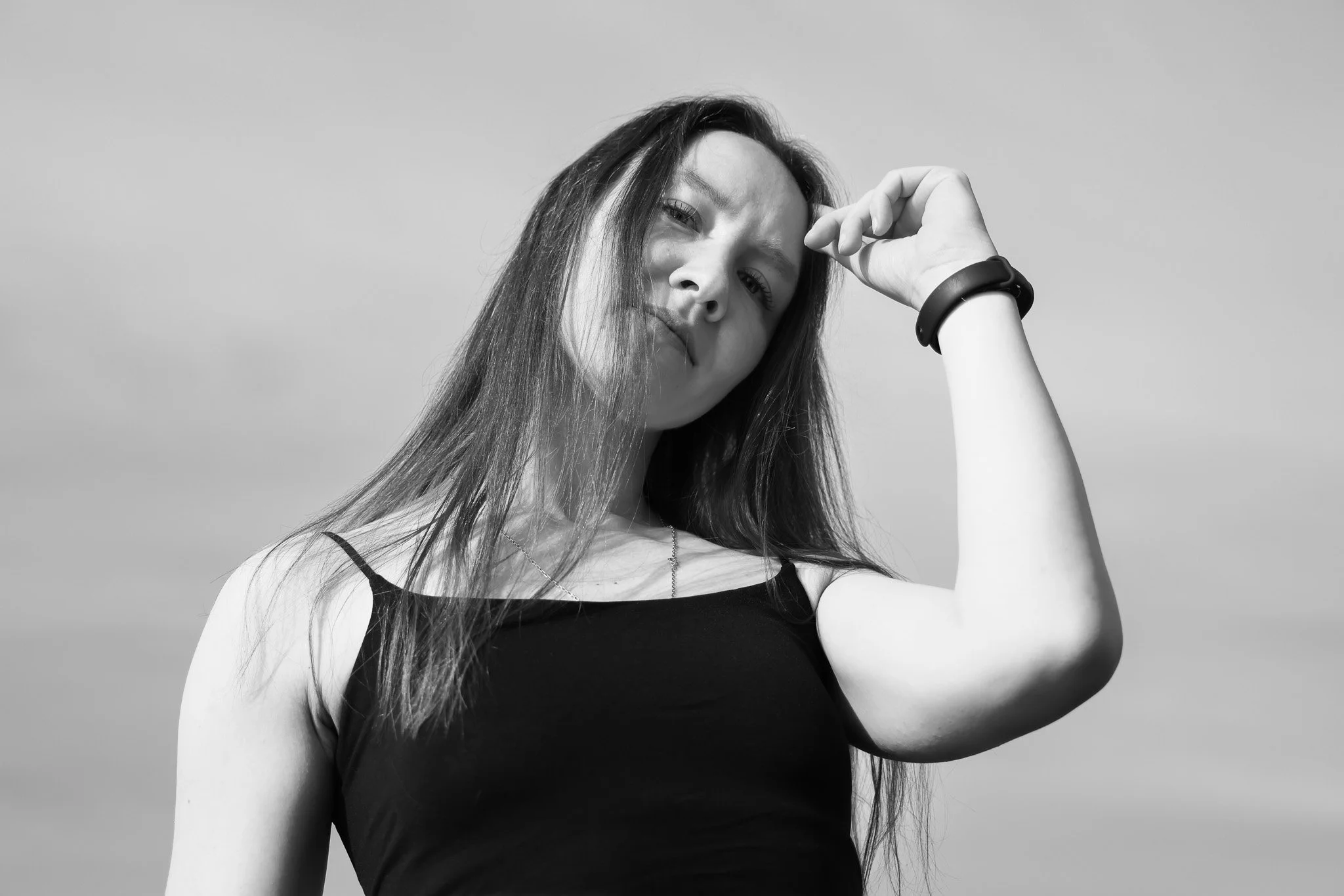 A young woman with long hair wearing a black tank top and a wristband, posing outdoors against a clear sky, looking at the camera with a slight tilt of her head.
