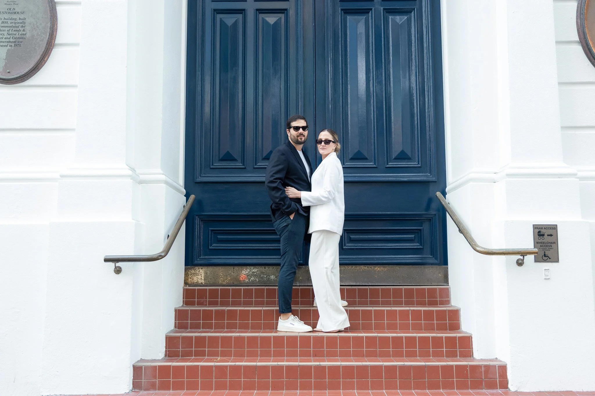 A man and woman wearing sunglasses and stylish clothes stand closely together on red tiled steps in front of a large blue door. The woman is dressed in white, and the man in black, with modern urban surroundings.