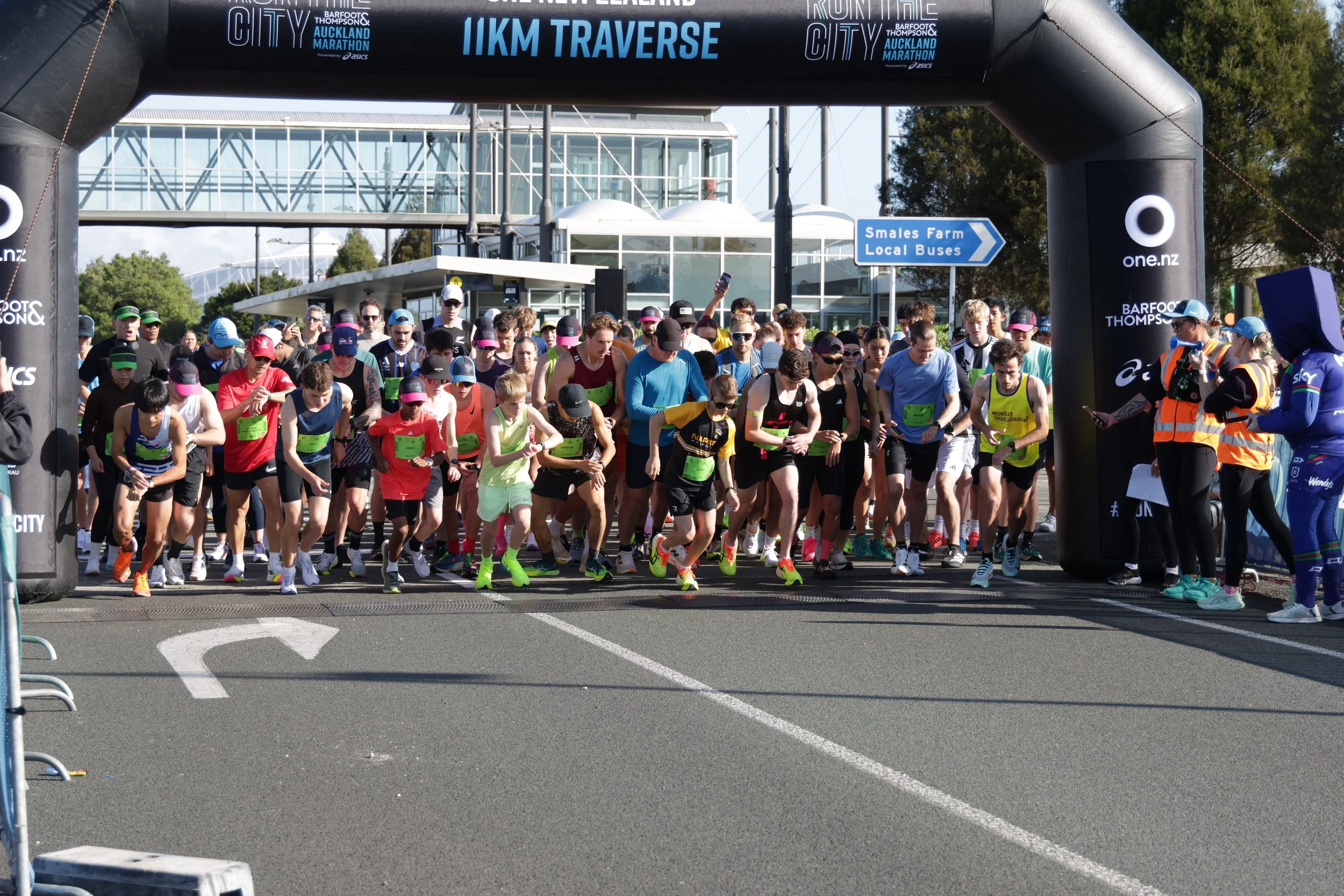 A large group of runners at the starting line of a marathon race, gathered under an archway with race banners and a modern building in the background.