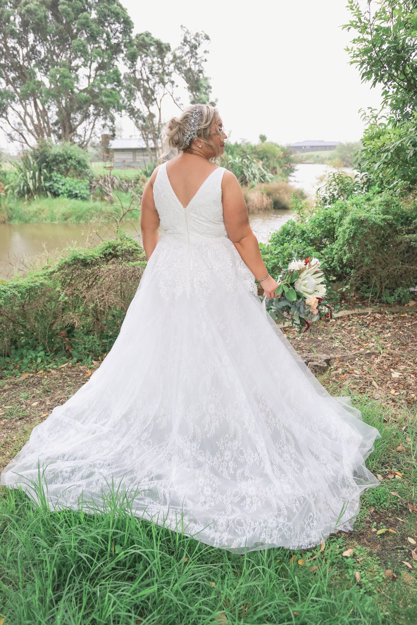 A bride wearing a white wedding dress holding a bouquet standing outdoors by a river with greenery and trees in the background.