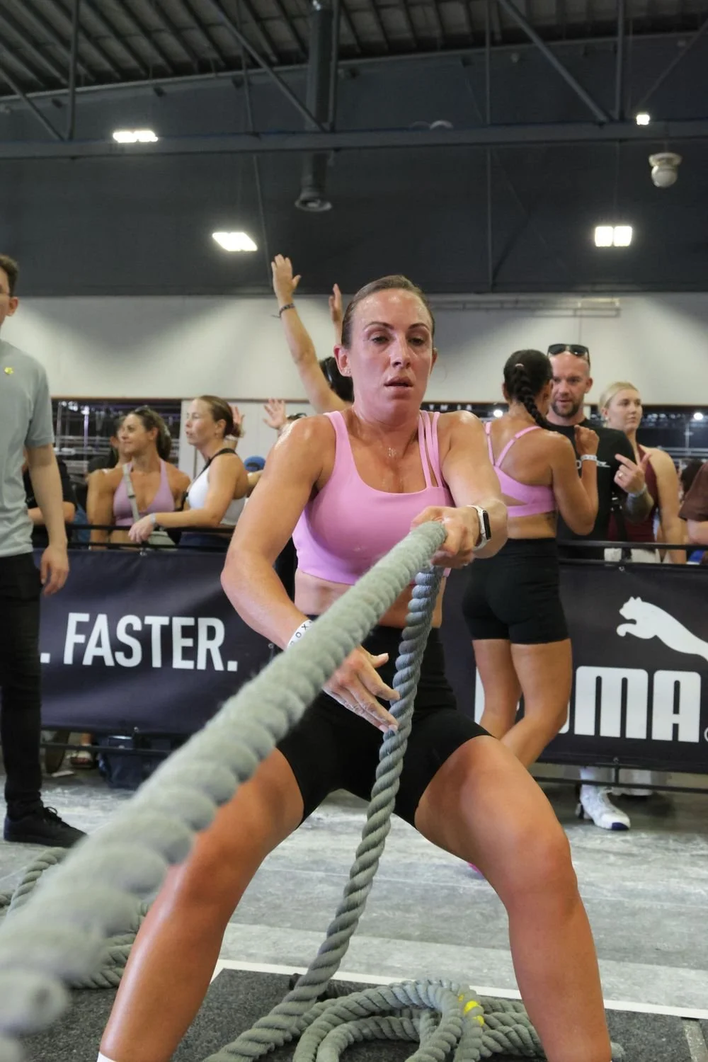 A woman in a pink sports bra and black shorts is engaged in a strong battle of tug-of-war with thick ropes during a fitness competition, with onlookers and event banners in the background.