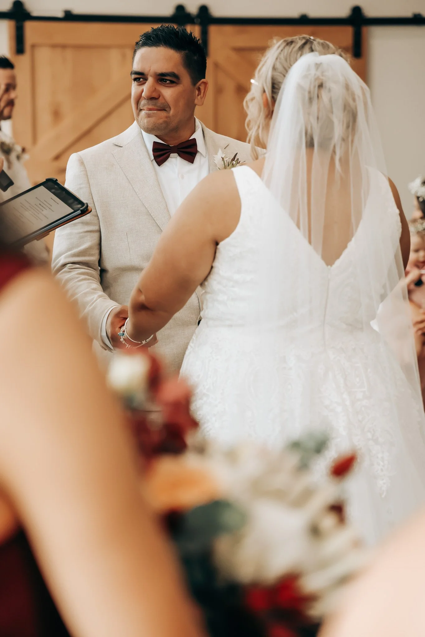 A man in a beige suit with a burgundy bowtie is looking on emotional as he holds hands with a woman in a white wedding dress and veil during a wedding ceremony inside a rustic wooden venue.