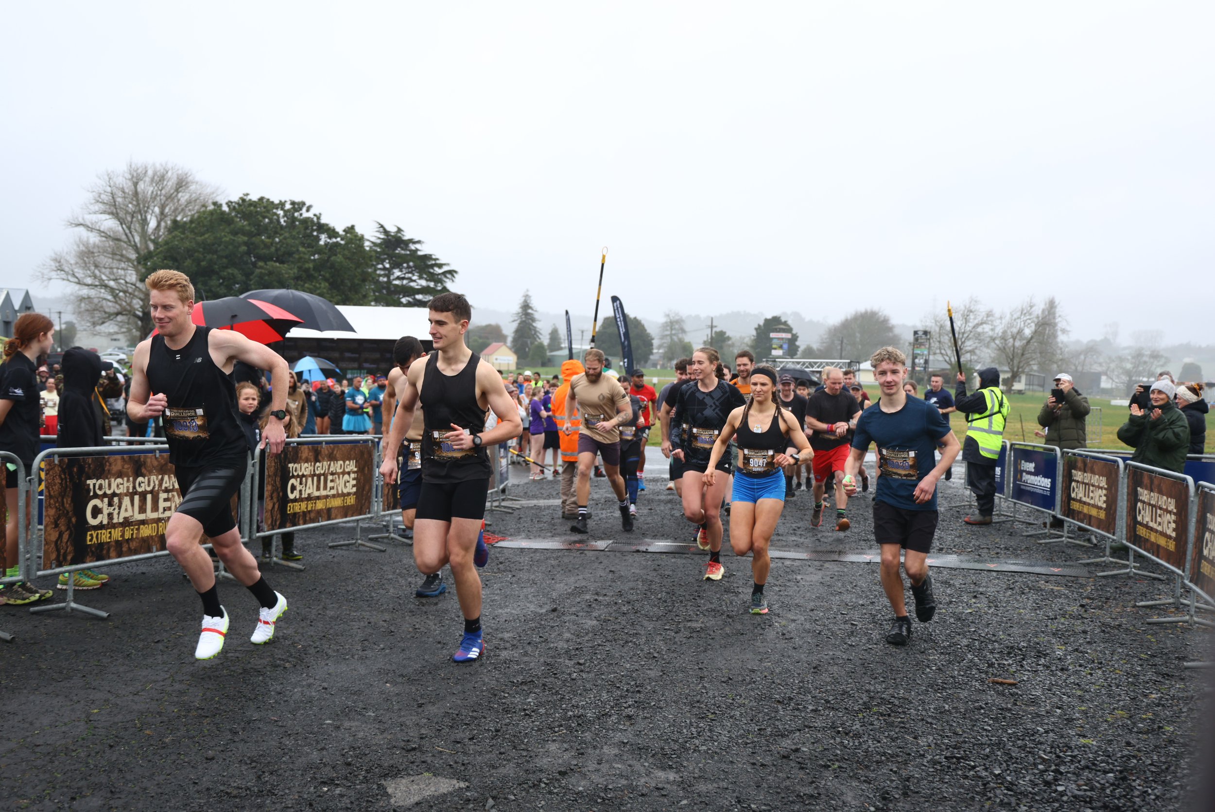 Runners crossing a finish line at an outdoor race event despite rainy weather, with spectators and event staff observing.
