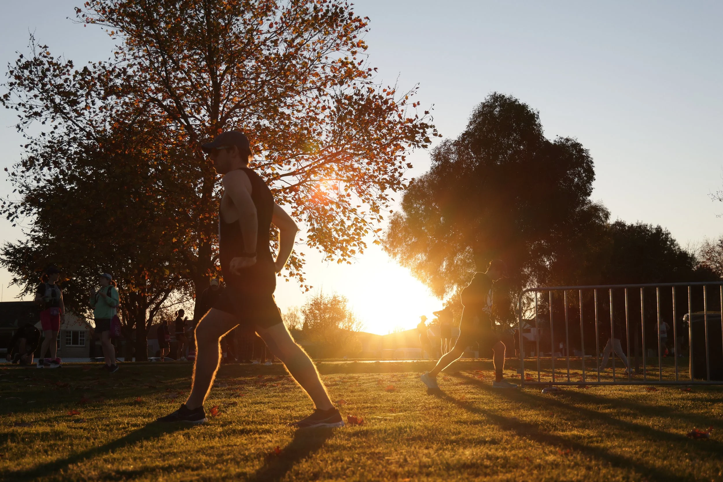 People jogging and socializing in a park at sunset, with trees displaying autumn foliage.