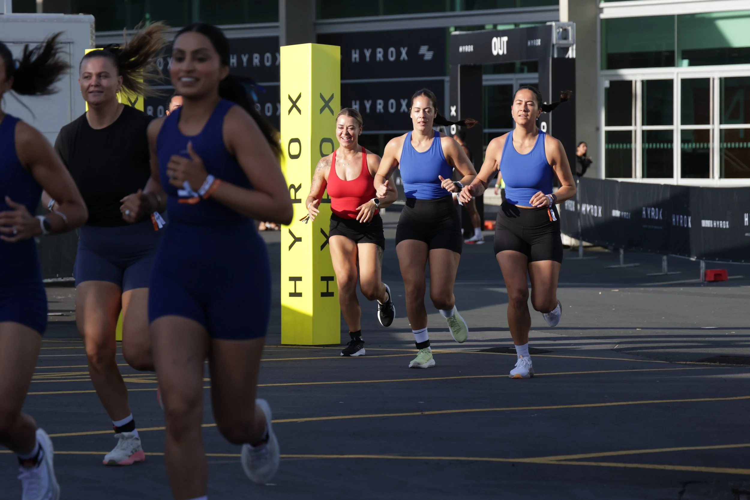 A group of women running in a race, wearing athletic clothing, on a track with a yellow timing post labeled 'HYROX' in the background.
