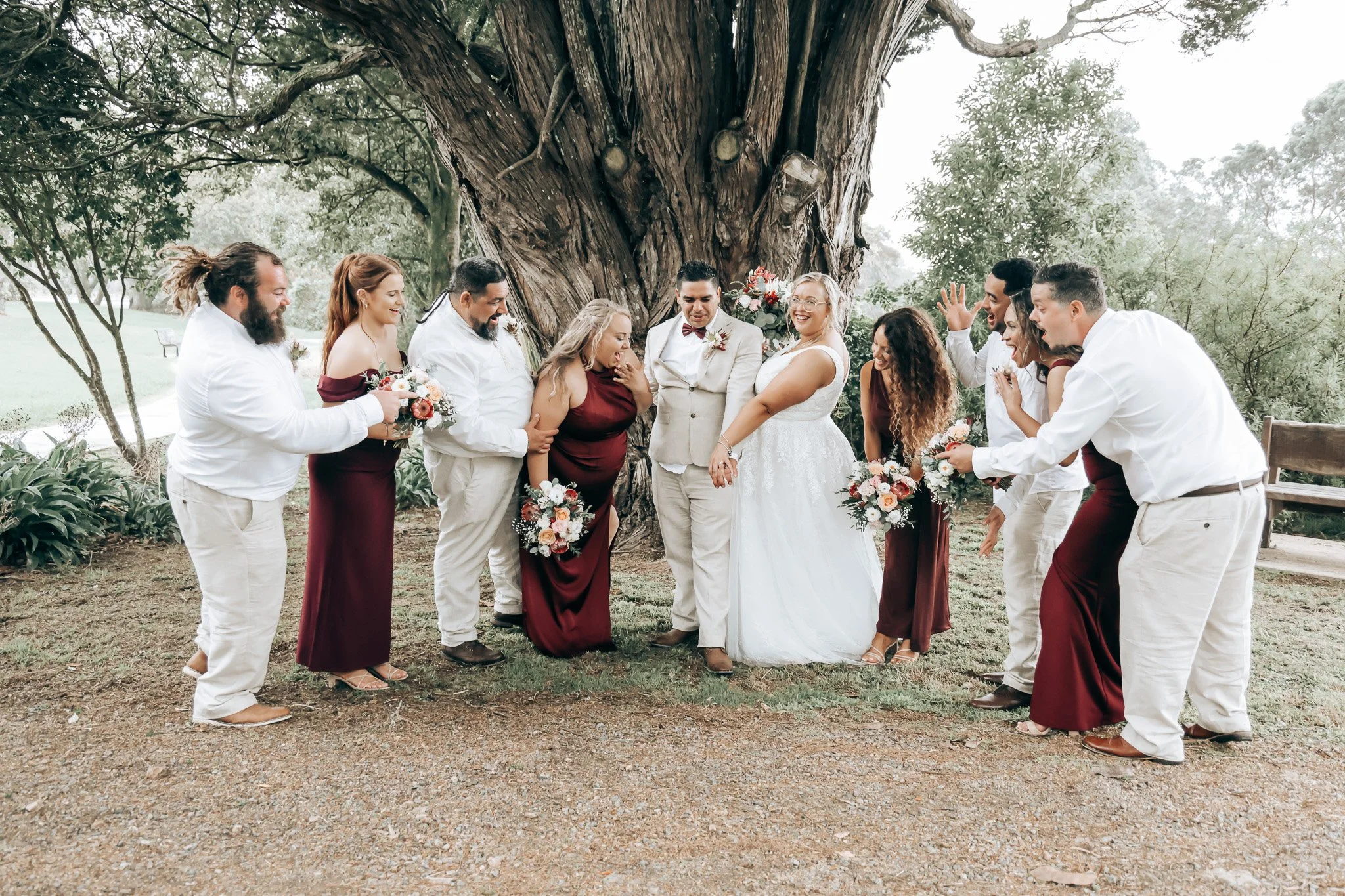 A group of people dressed in wedding attire, standing outdoors in front of a large tree, celebrating at a wedding ceremony.