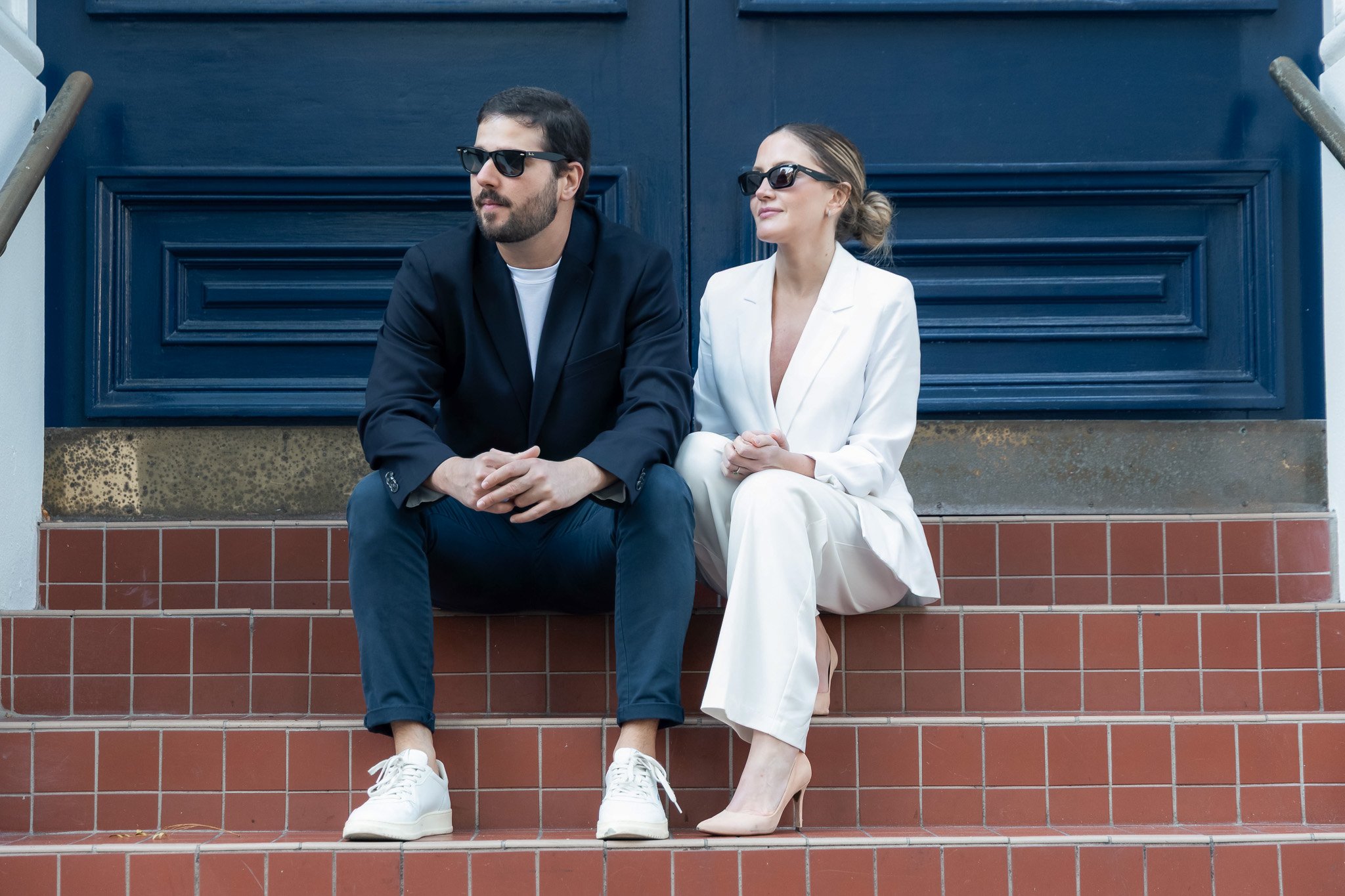 A man and woman sitting on brick steps in front of a dark blue door, both wearing sunglasses. The man is dressed in a black blazer, white T-shirt, and blue jeans, with white sneakers. The woman is wearing a white suit, with her hair tied back and bei