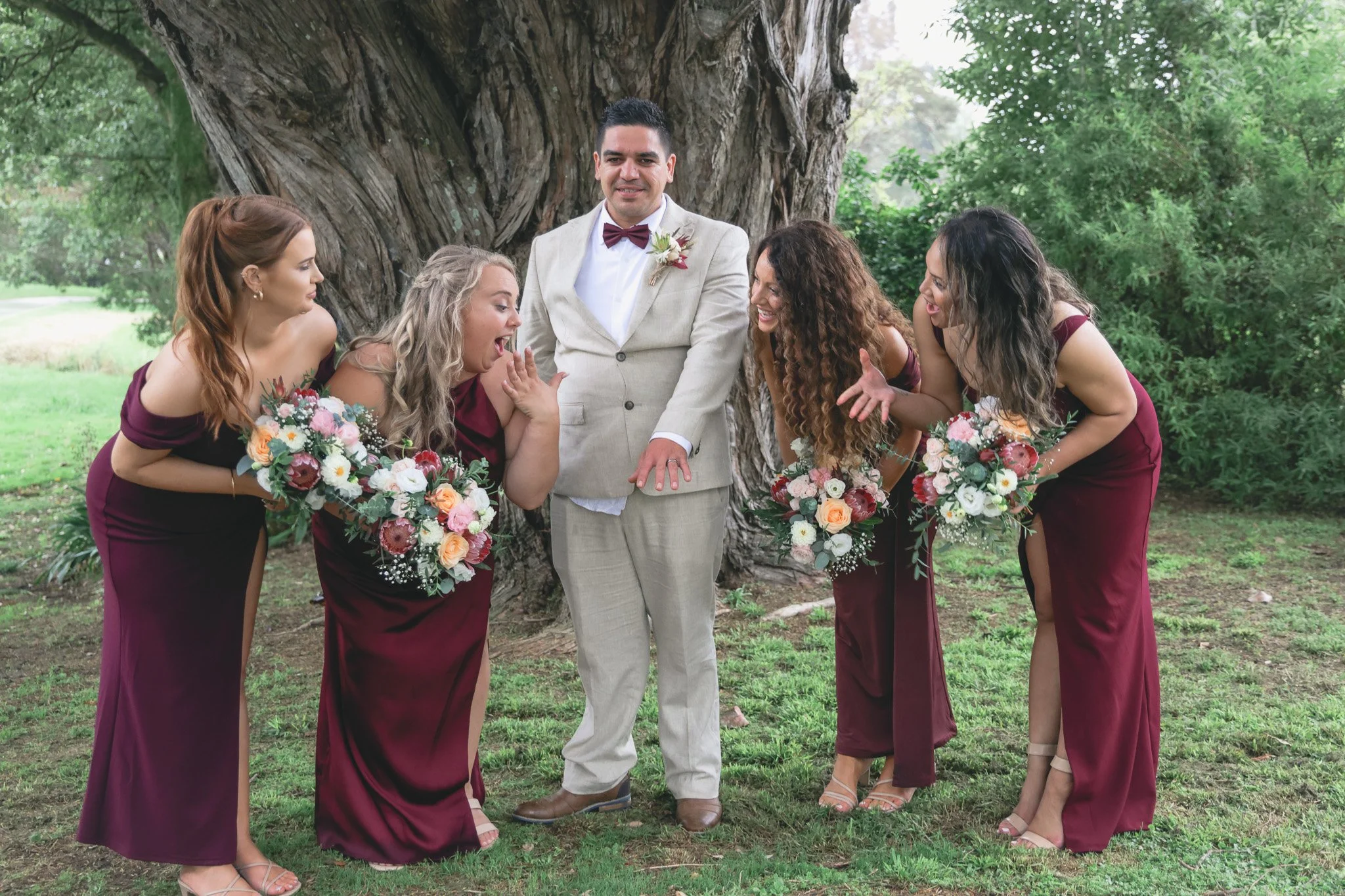 A groom in a beige suit with a maroon bow tie and boutonniere stands under a large tree, surrounded by four bridesmaids in burgundy dresses holding bouquets, all outdoors with green foliage.