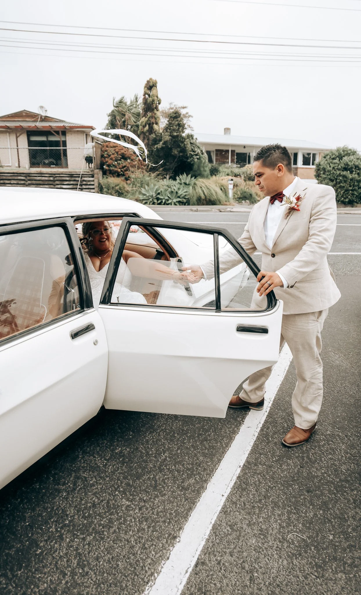 A groom holding hands with a bride sitting inside a white car during a wedding, standing outside on a parking lot, with houses and trees in the background.