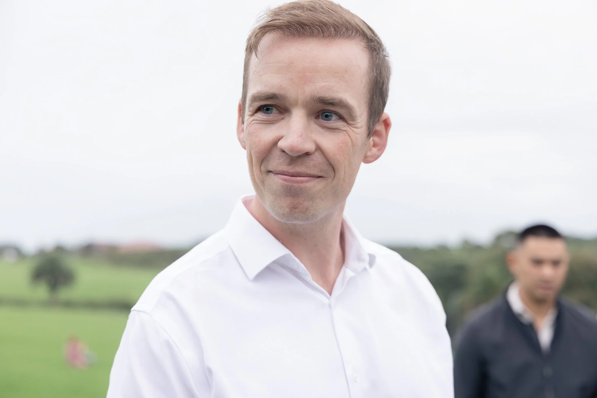 A man with short brown hair wearing a white shirt, smiling slightly, outdoors on a cloudy day, with other people in the background.