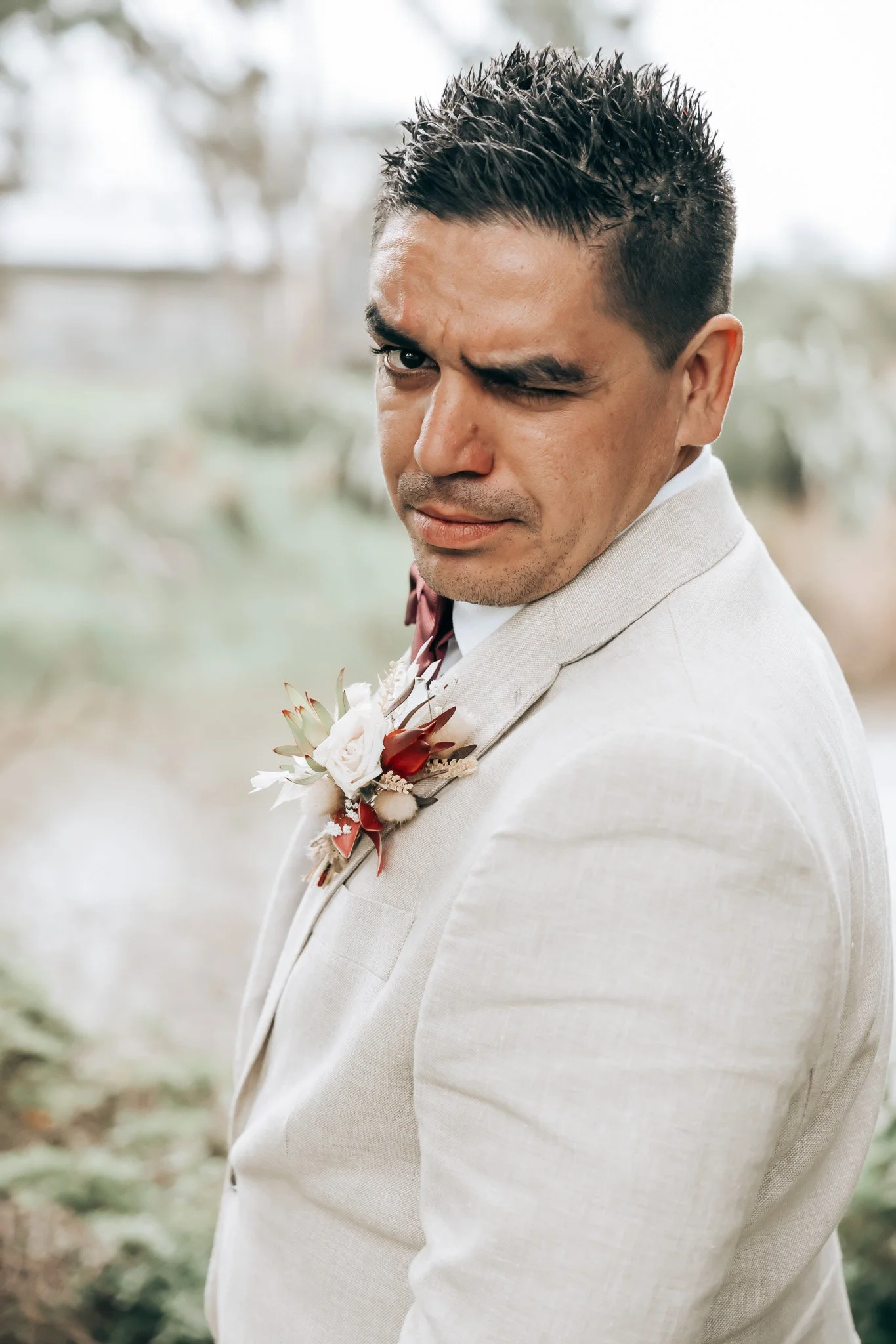 A man in a light-colored suit and a floral boutonniere, making a playful facial expression while winking at the camera outdoors.