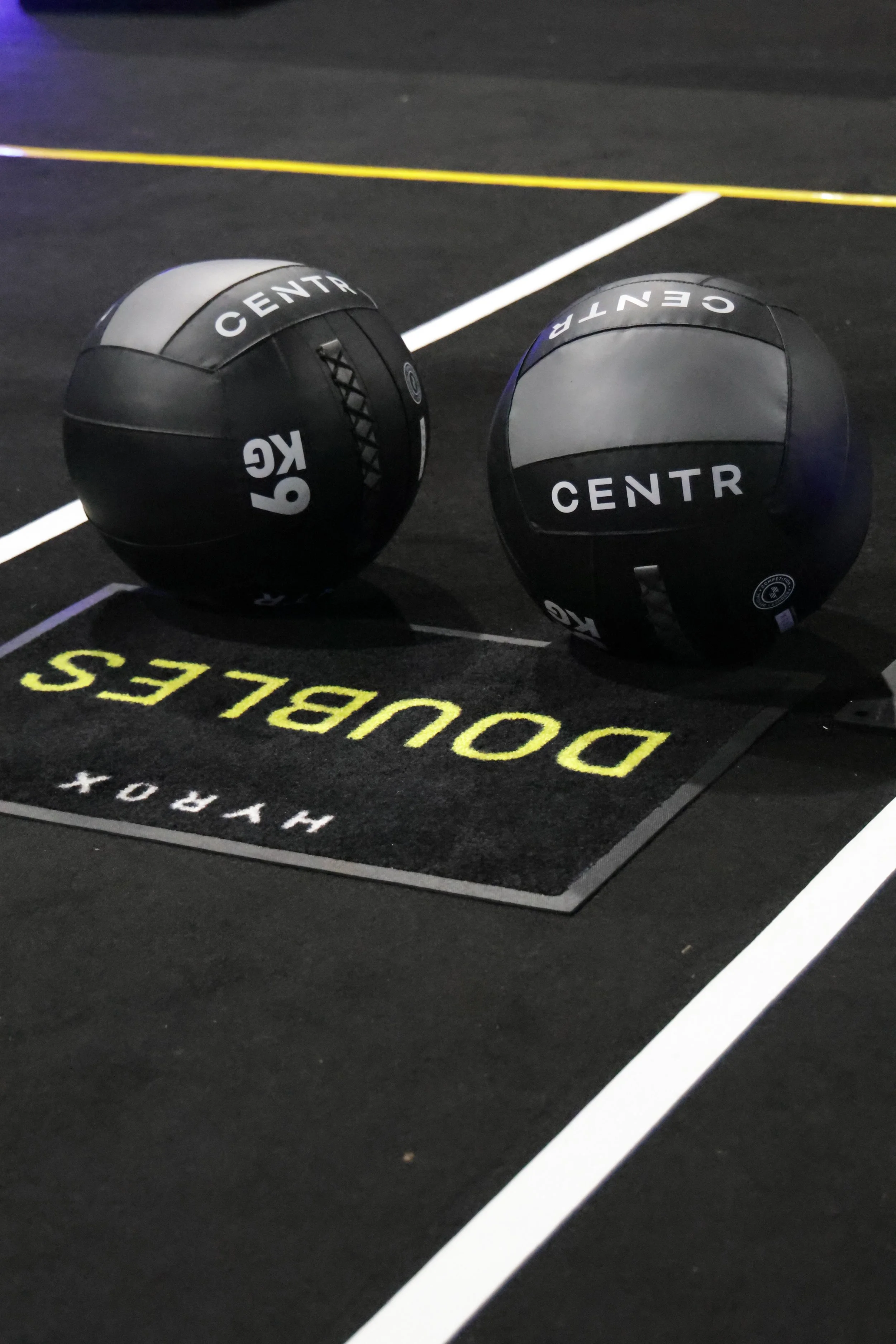 Two black medicine balls with white and gray markings resting on a black gym floor with white and yellow lines and text.