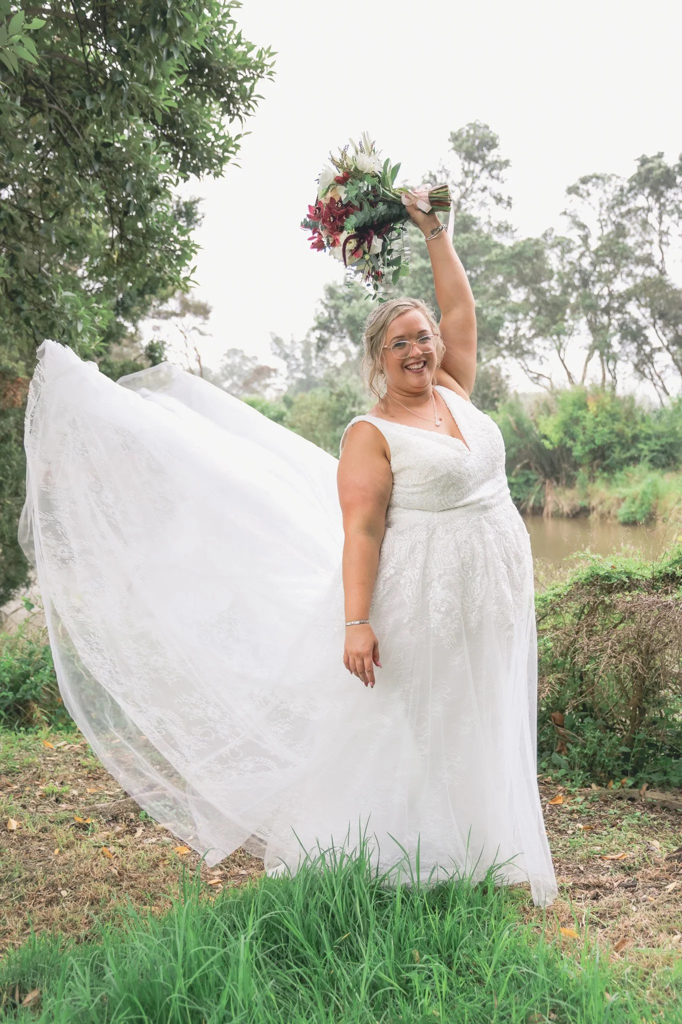 A woman in a white wedding dress holding a bouquet of flowers in the air outdoors near a small river, with greenery and trees in the background.