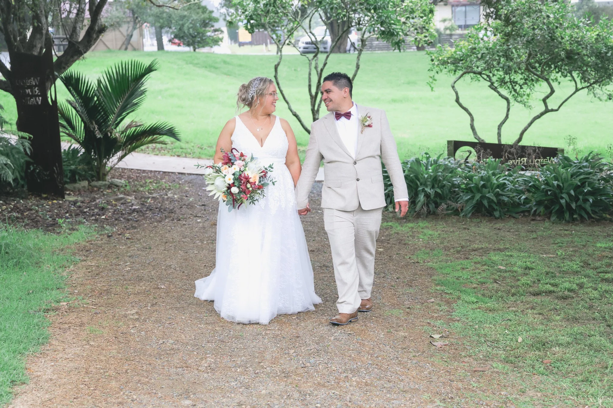 A bride and groom walking outdoors hand in hand, smiling and looking at each other in a park with green grass and trees.