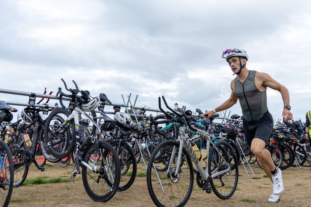 A man with a helmet and sleeveless athletic top is running with a bicycle among many parked bikes at a triathlon transition area.
