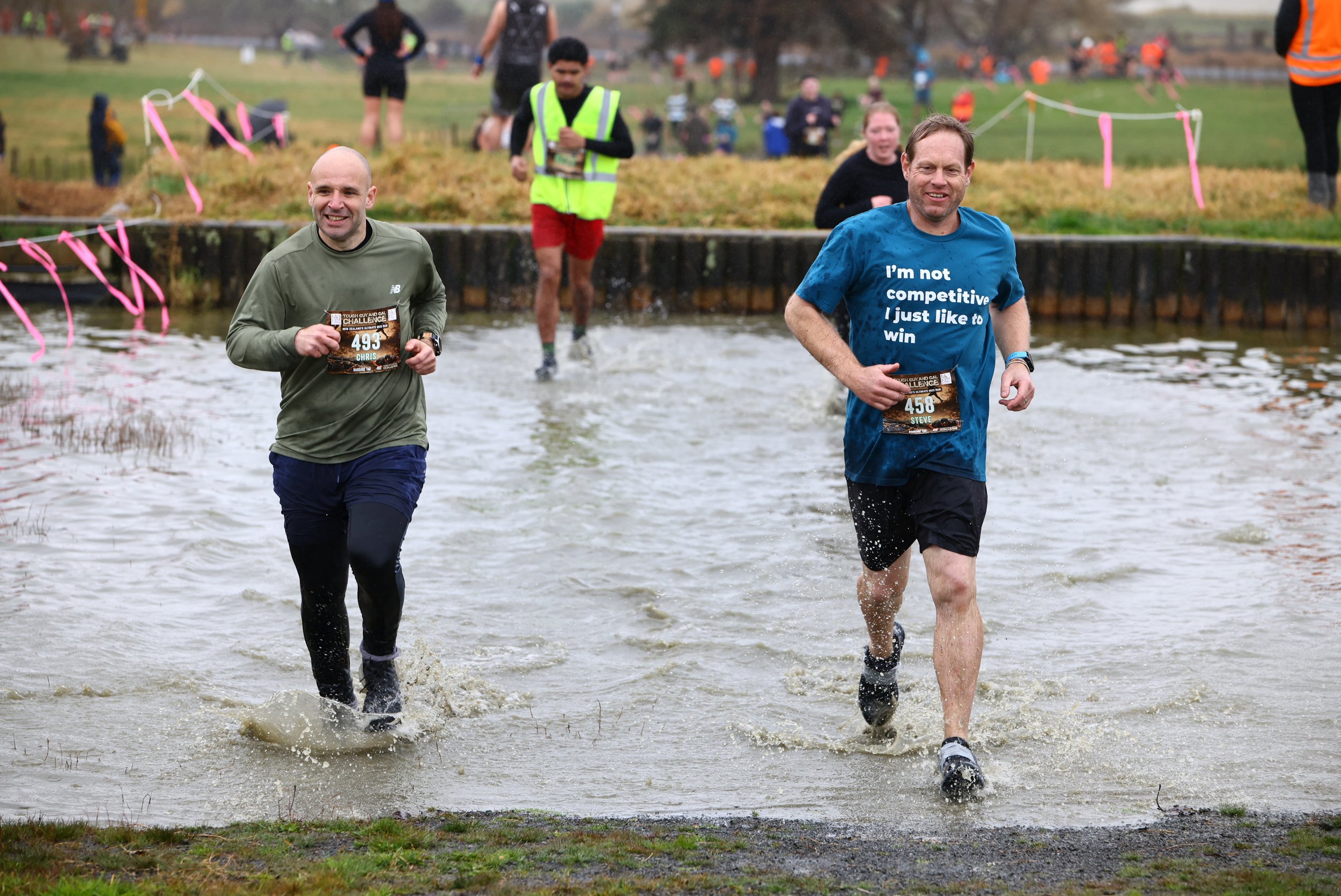 Two men running through a water obstacle during a race, with other runners and spectators in the background.