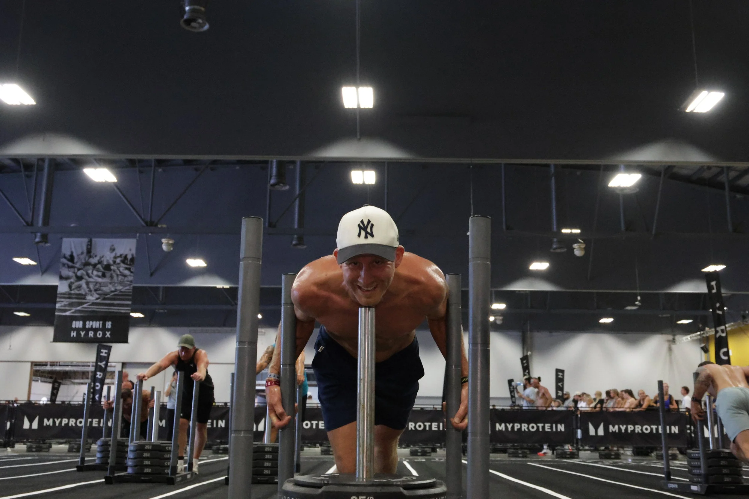 A man smiling and exercising at a fitness gym, with other people working out in the background.