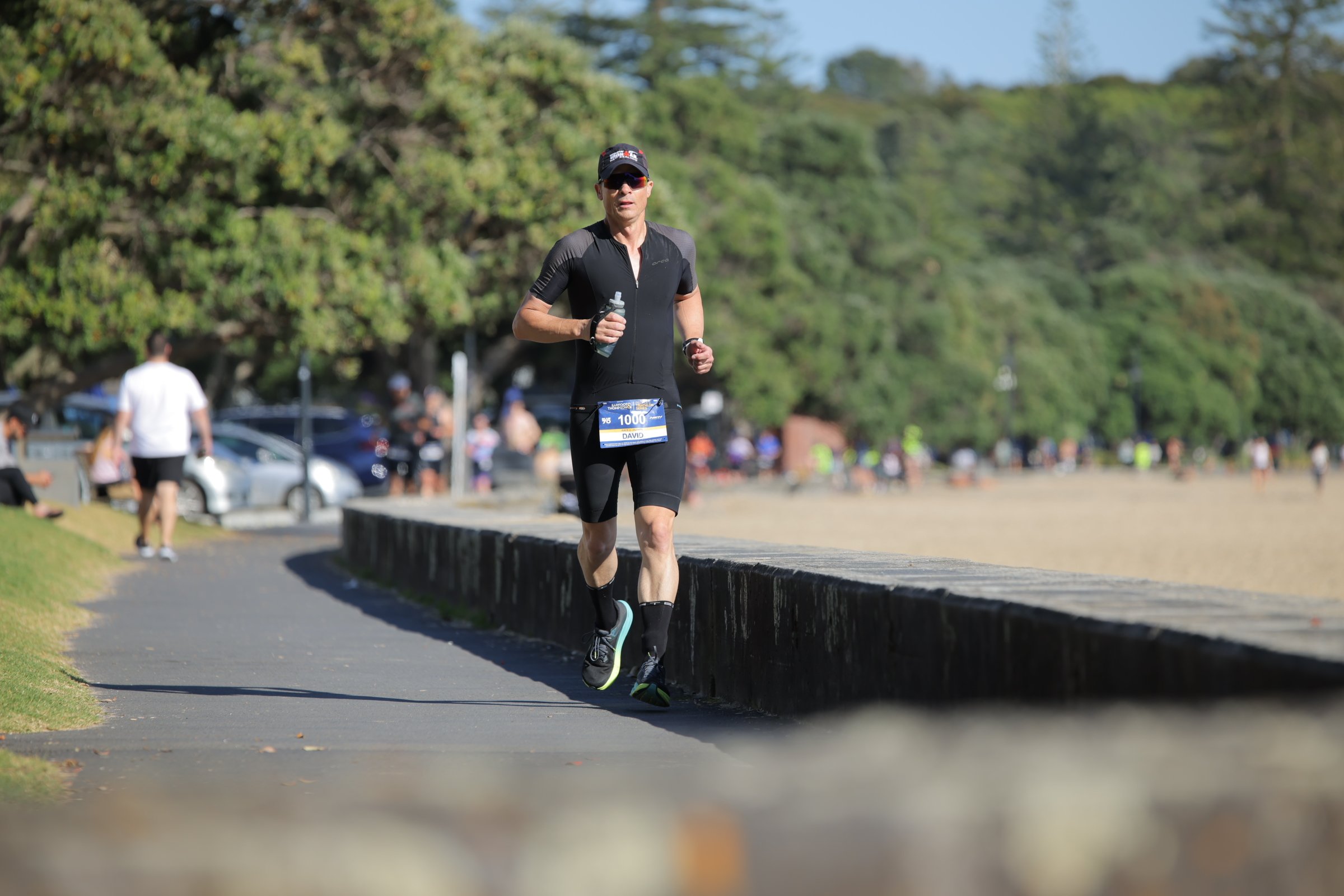 A man running outdoors on a paved path during a race, wearing athletic clothing, sunglasses, a cap, and a race bib with the number 1000.