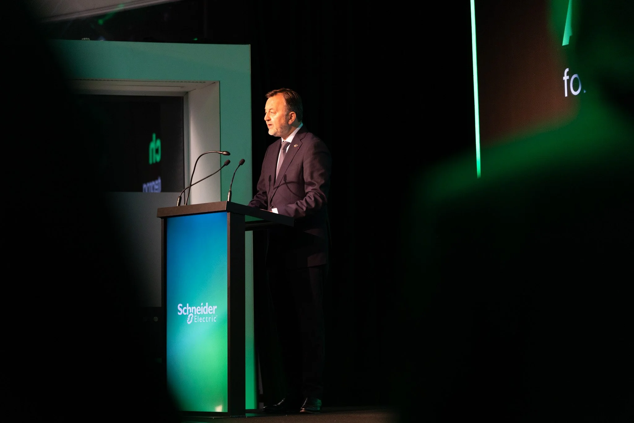 A man in a suit standing at a podium with microphones, delivering a speech at a conference or event. The background has a screen displaying the Schneider Electric logo and branding.