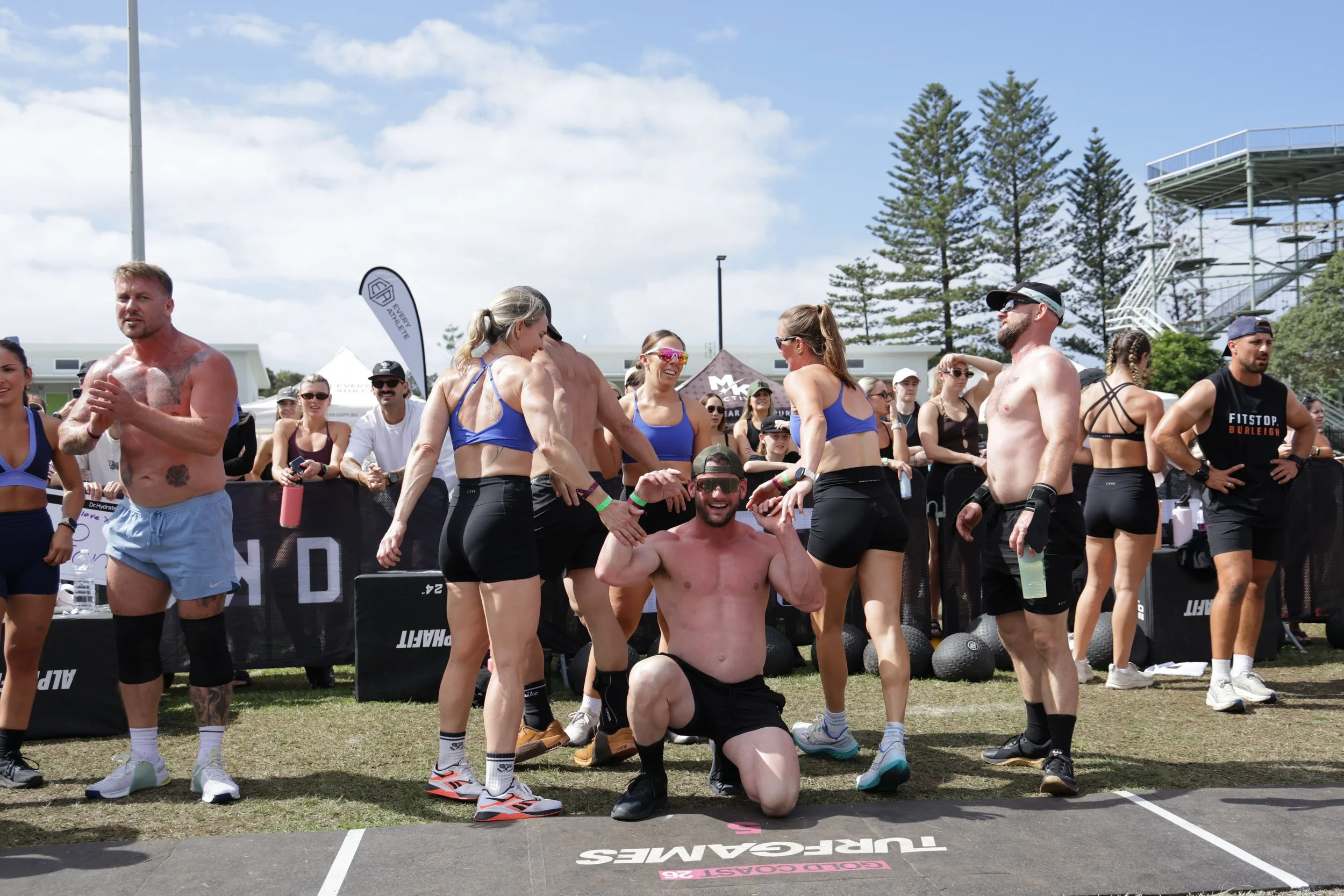 Group of fitness participants and spectators outdoors at a CrossFit event, with some people shirtless, in athletic gear, and smiling.