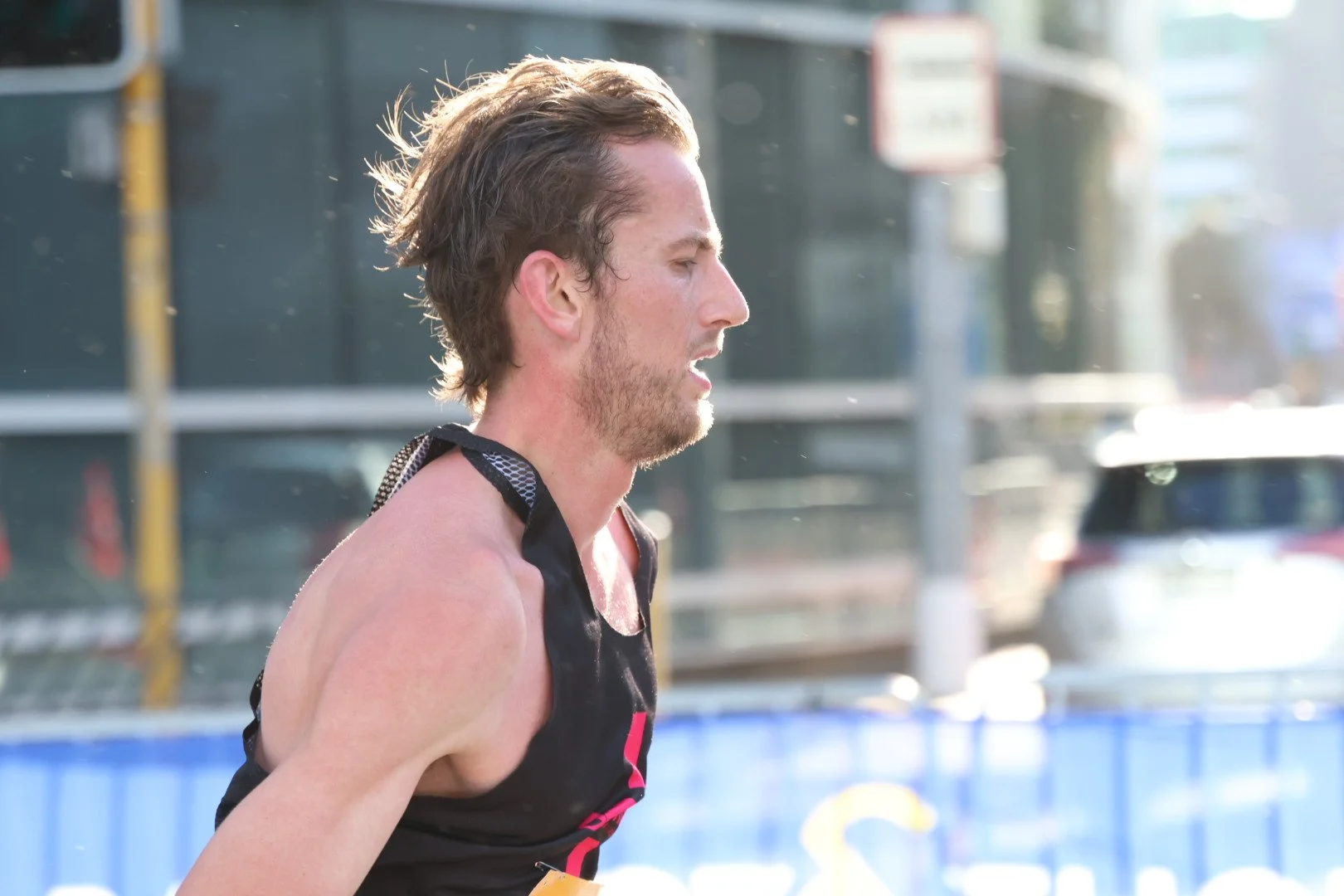 A male runner with short brown hair and facial hair in profile, wearing a black sleeveless athletic shirt, running outdoors on a track during daytime.