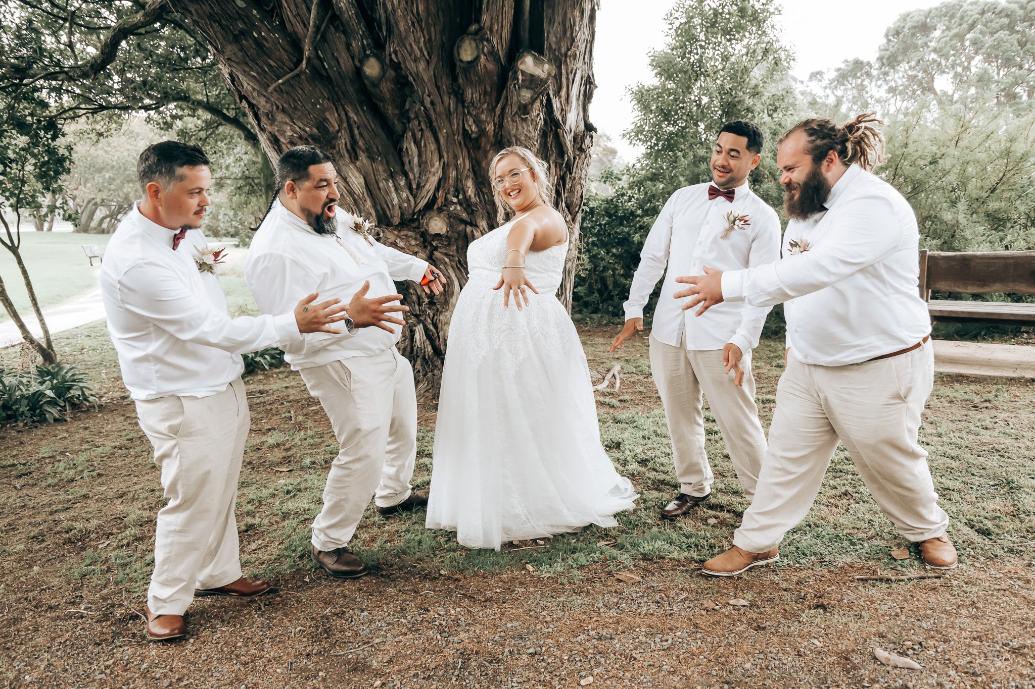 A group of five men and one woman celebrating outdoors near a large tree during a wedding. The woman is in a wedding dress and the men are in white shirts and beige pants, with some wearing bow ties and boutonnieres. They are all smiling and appear t