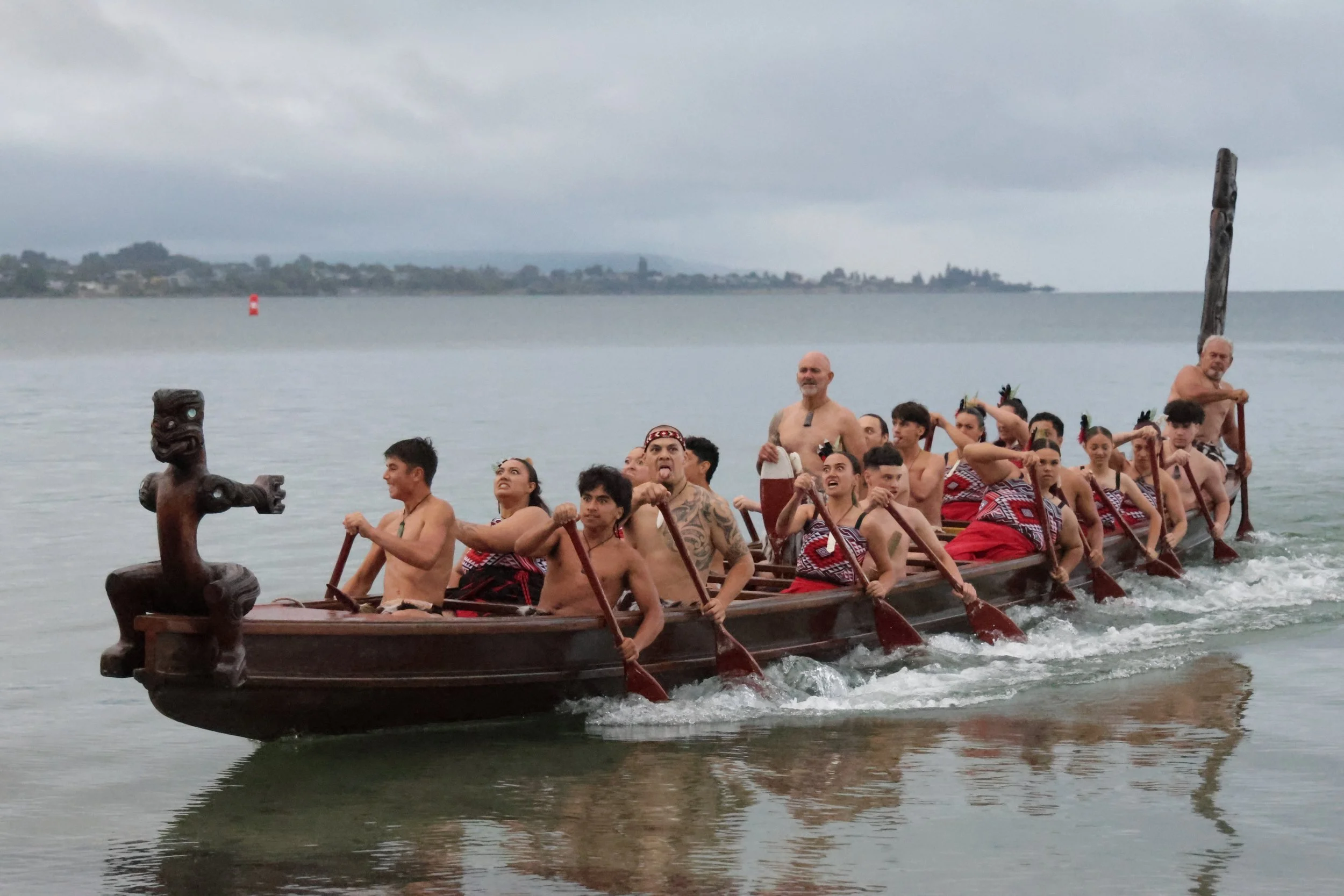 A group of people on a traditional Polynesian outrigger canoe, paddling on calm water during overcast weather, with a shoreline and cloudy sky in the background.