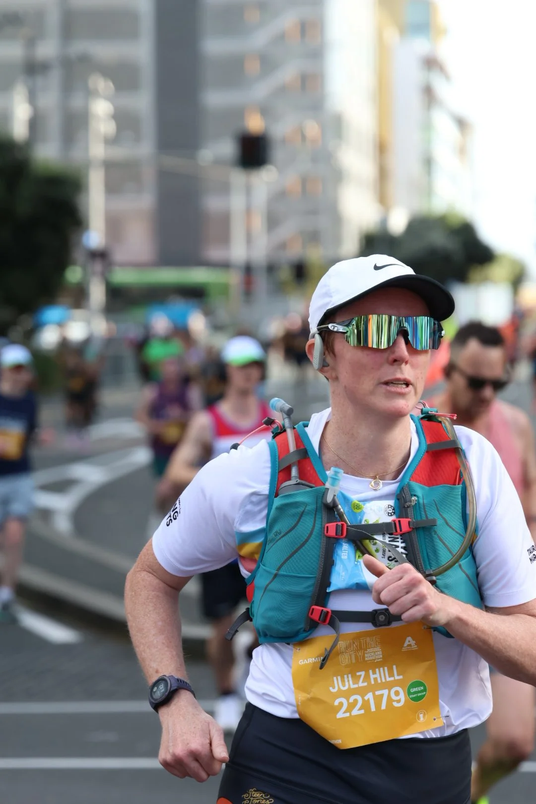 A woman participating in a marathon, wearing sunglasses, a white Nike cap, and a running vest with a race bib that reads 'Julz Hill 22179'. She is surrounded by other runners on a city street.
