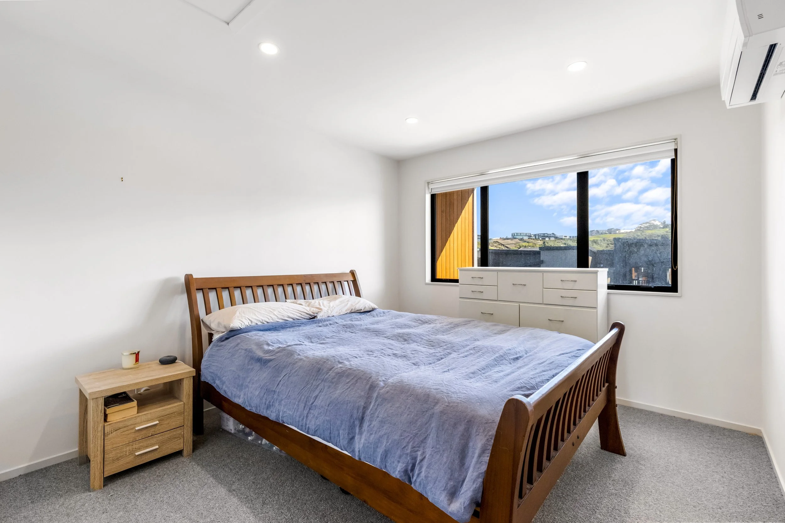 A bedroom with a wooden bed frame, a blue bedspread, a window showing outside scenery, a white dresser, a small nightstand with a candle and remote, gray carpet, white walls, ceiling lights, and an air conditioning unit.