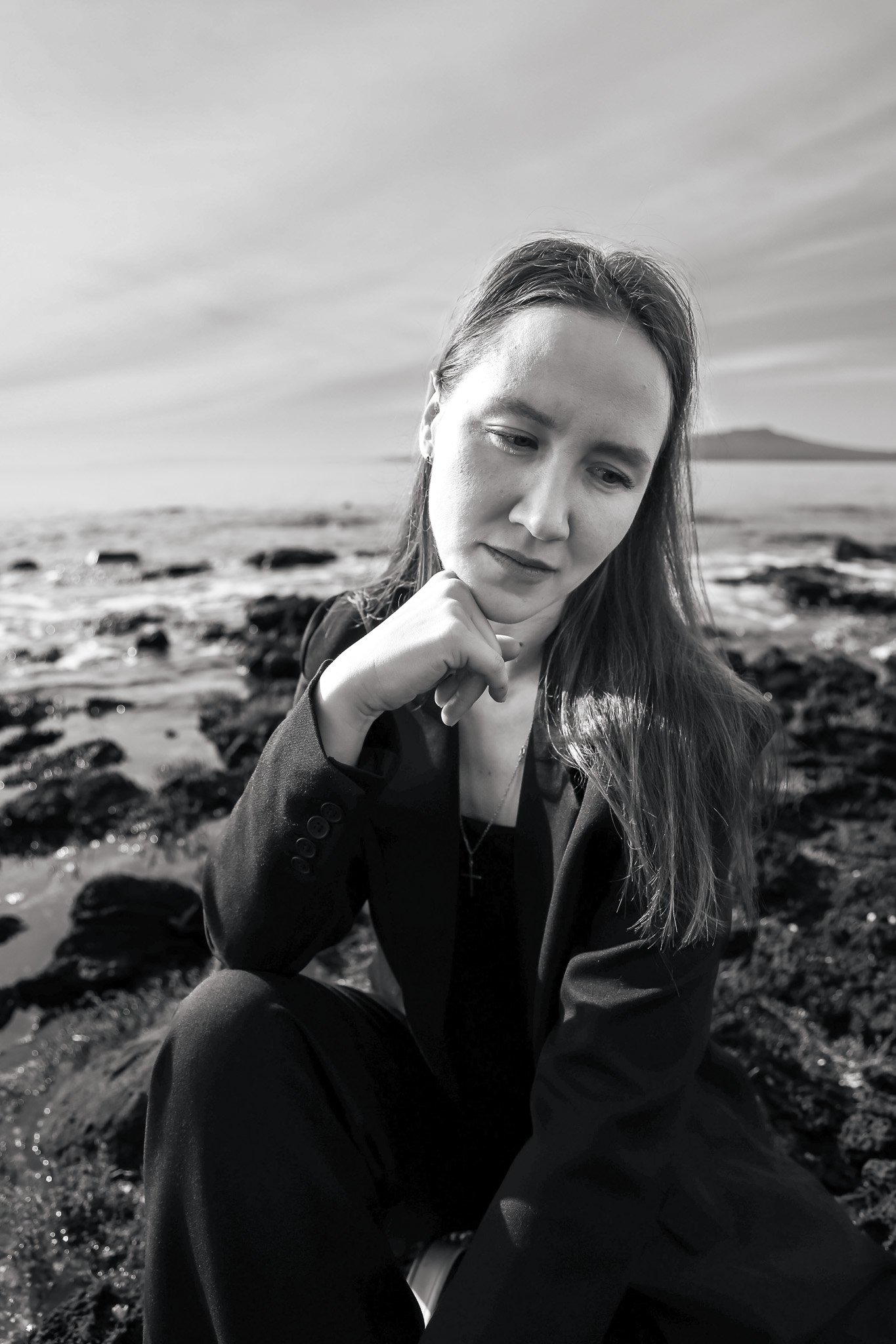 A woman with long hair and light skin sitting outdoors on rocky terrain, looking thoughtful, dressed in dark clothing, with a body of water and distant land in the background, under a cloudy sky, in black and white.