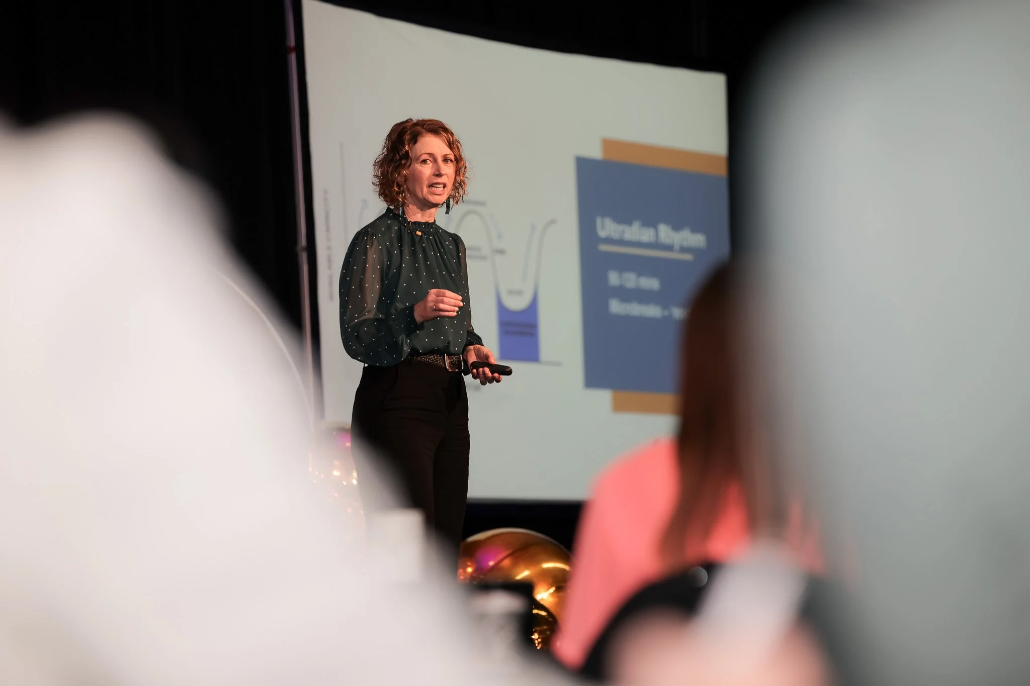 A woman with curly hair giving a presentation in front of a large screen, with audience members' heads visible in the foreground.