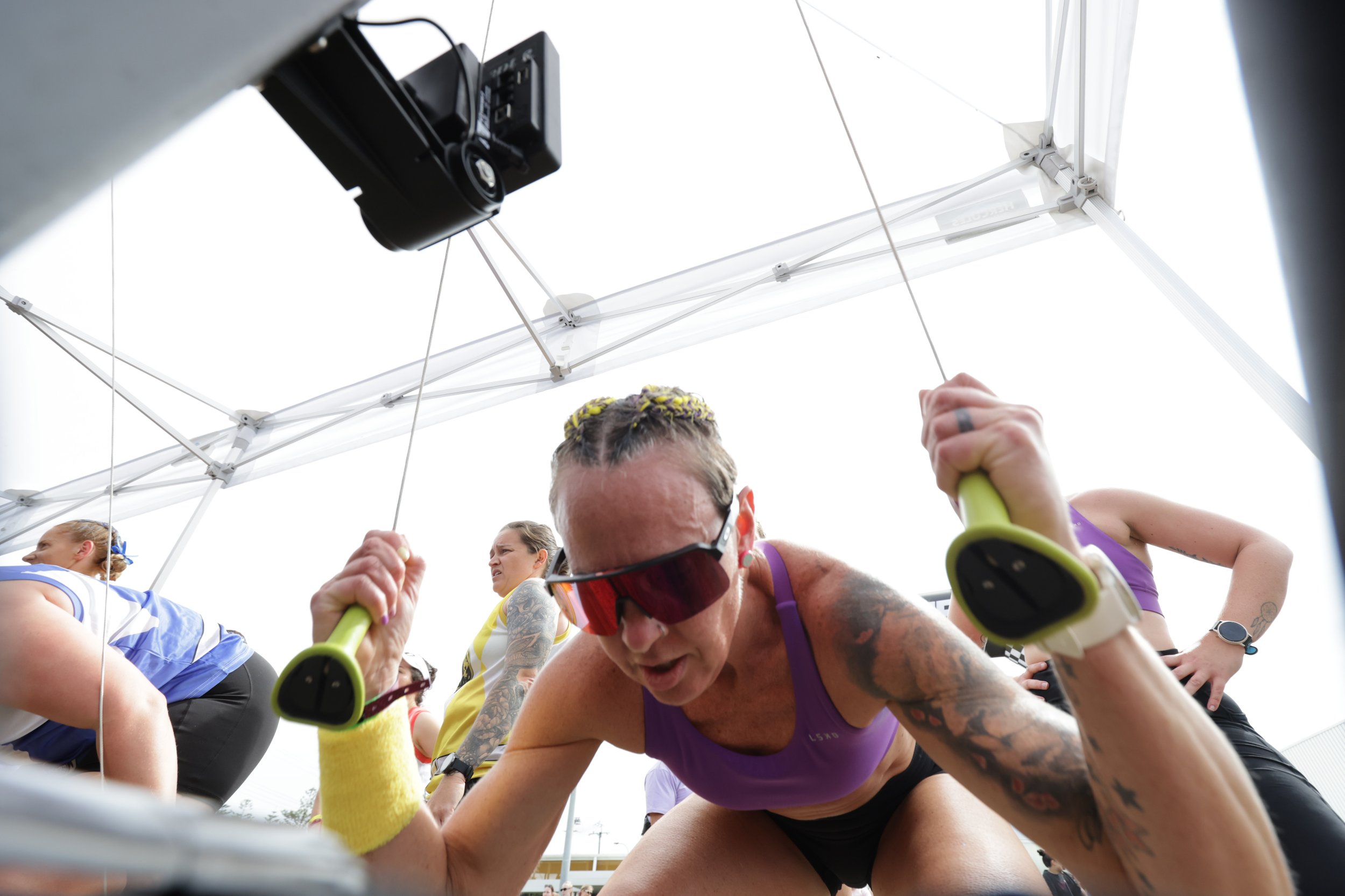 A group of runners participating in a race, seen from below through a gap in the starting block or platform. They are wearing athletic clothing and some have tattoos. The sky is cloudy.
