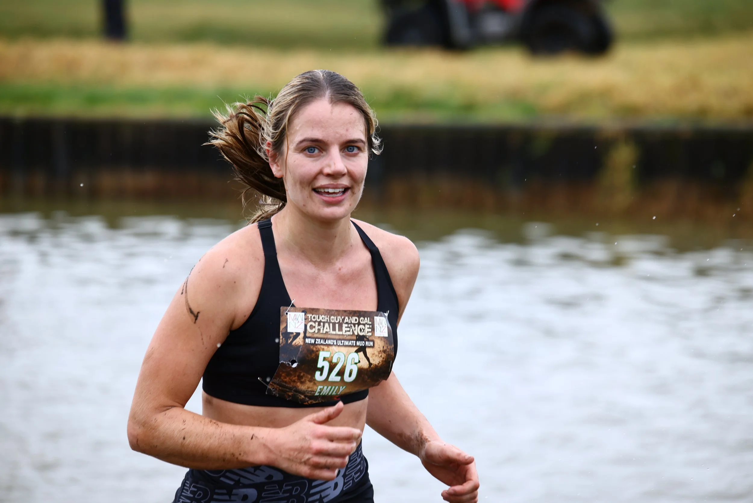 A young woman with wet hair running in a muddy outdoor race, wearing a black sports bra and shorts, with a bib number 526 reading "Emily," participating in a challenging mud run event.