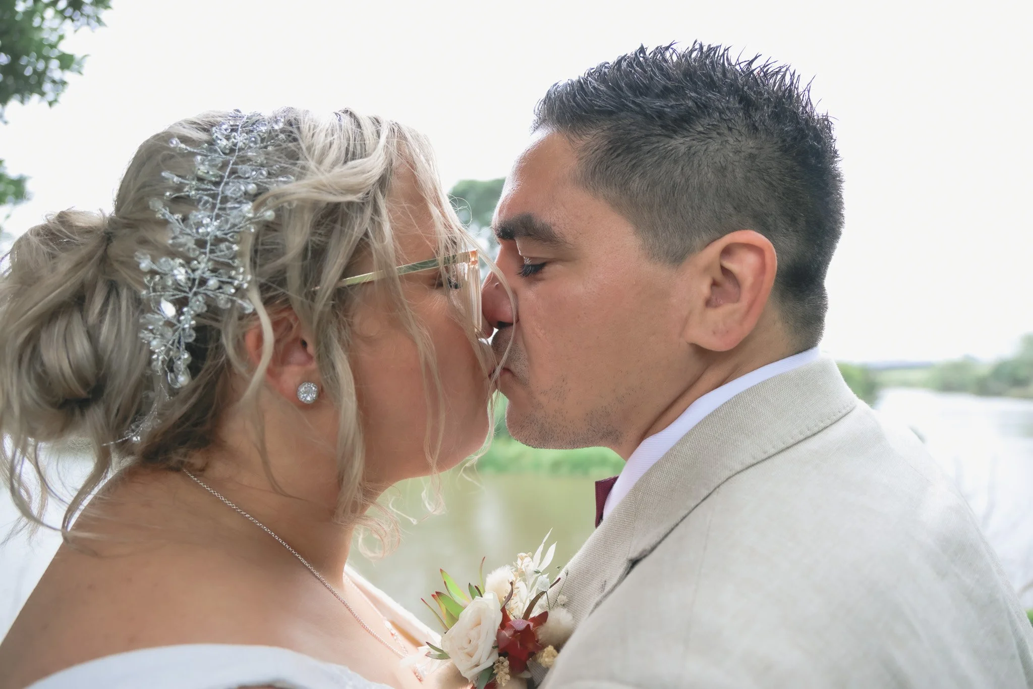 A bride and groom are about to kiss during their wedding ceremony outdoors. The bride has curly blonde hair with a jeweled hairpiece and is wearing earrings. The groom has short, dark, spiked hair and is dressed in a light-colored suit with a white s