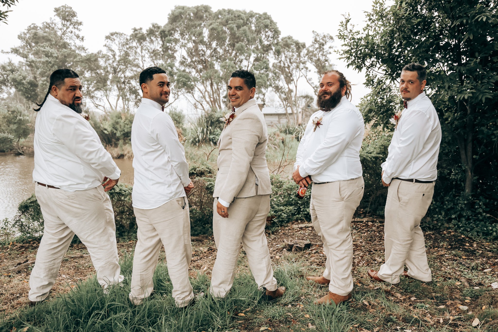 Group of five men dressed in white shirts and beige pants standing outdoors near a body of water, with trees in the background.