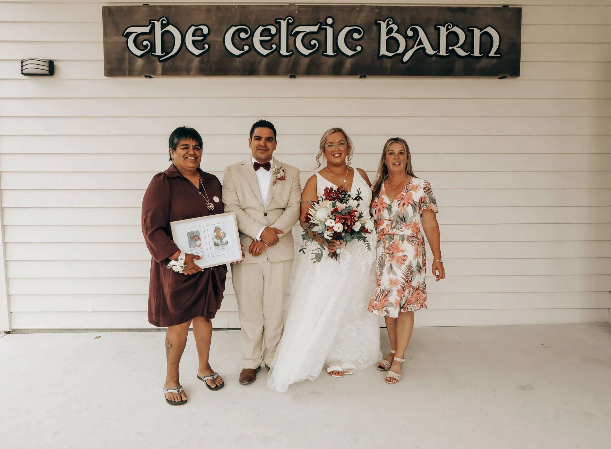 Four women and one man standing outside in front of a sign that reads "The Celtic Barn." The man and one woman are dressed in wedding attire, with the woman holding a bouquet of flowers, suggesting a wedding celebration.