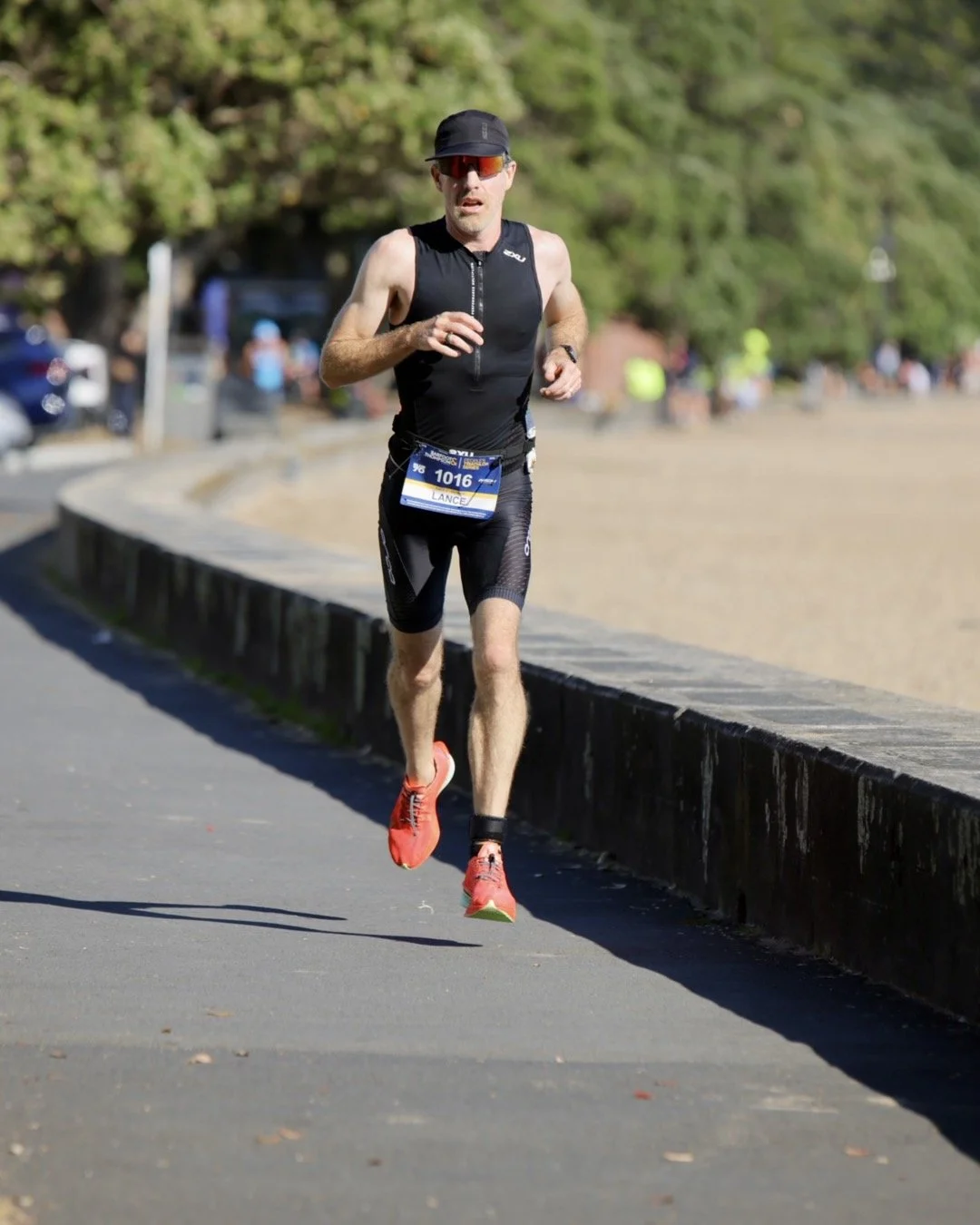 A male runner wearing a black athletic outfit, red running shoes, and sunglasses running along a paved path during a race, with trees and spectators in the background.