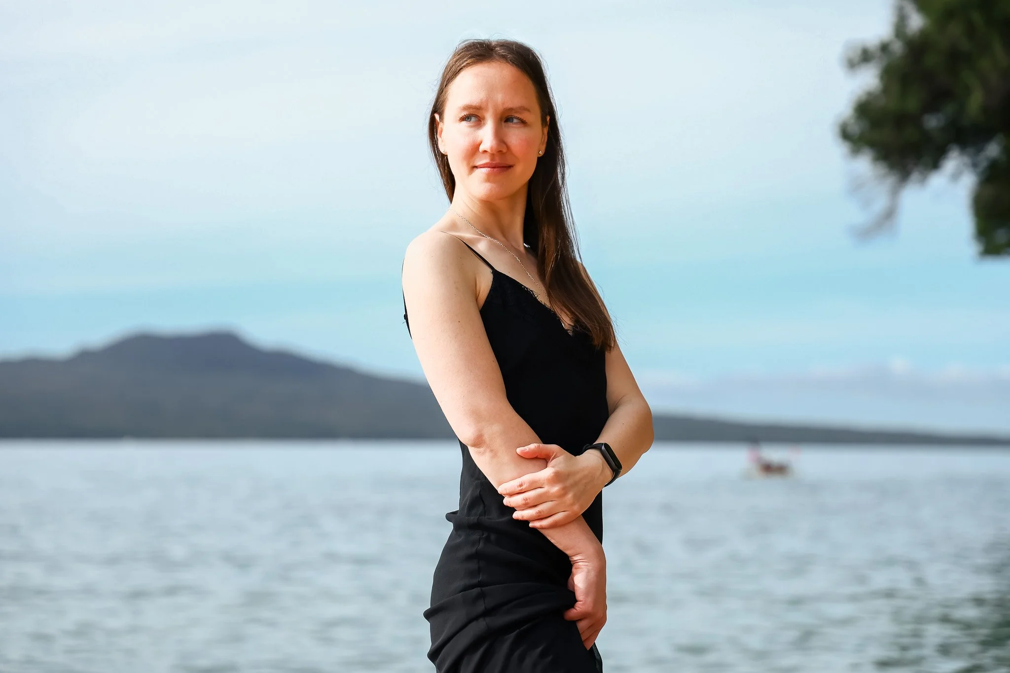 A woman with long brown hair in a black sleeveless top standing outdoors near a body of water with mountains in the background, looking to the side.