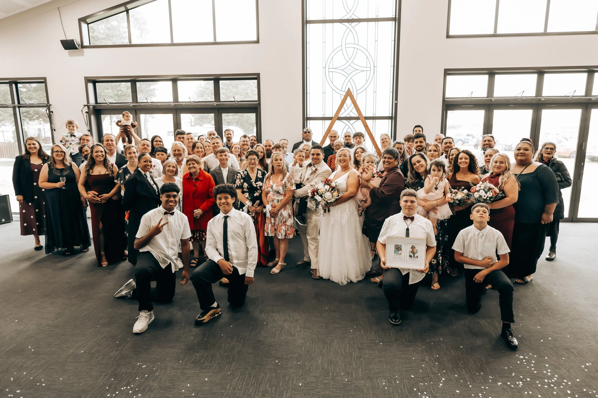 A large group of people, including children, teenagers, adults, and seniors, dressed semi-formally, gathered indoors in front of large windows for a celebration, with some holding flowers and a framed picture, with a stained glass design on the wall 