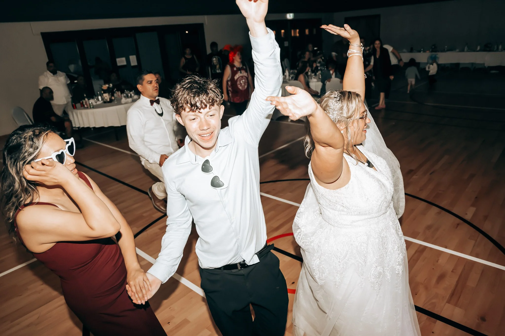 People at a wedding reception dancing on a wooden floor, with several guests in the background.