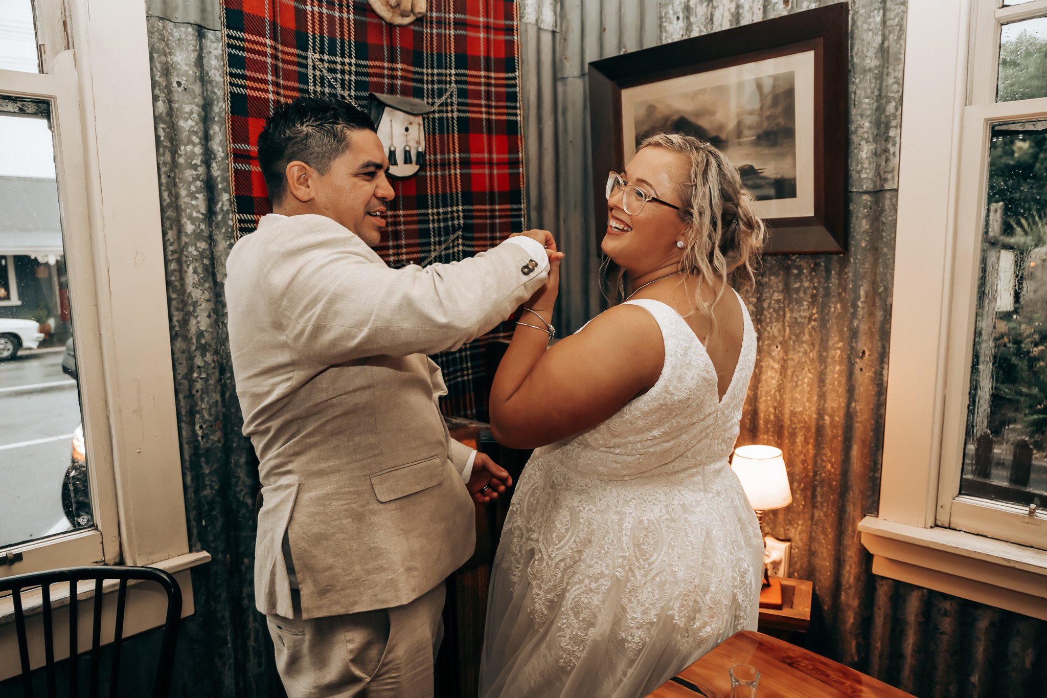 A couple is dancing and smiling in a cozy indoor setting with rustic walls, a framed picture, and a window showing rain outside.